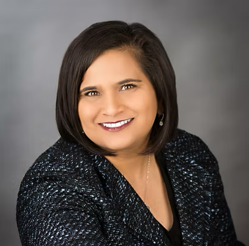 A professional woman with shoulder-length dark hair, smiling, wearing a black and blue textured jacket with earrings against a gray background.