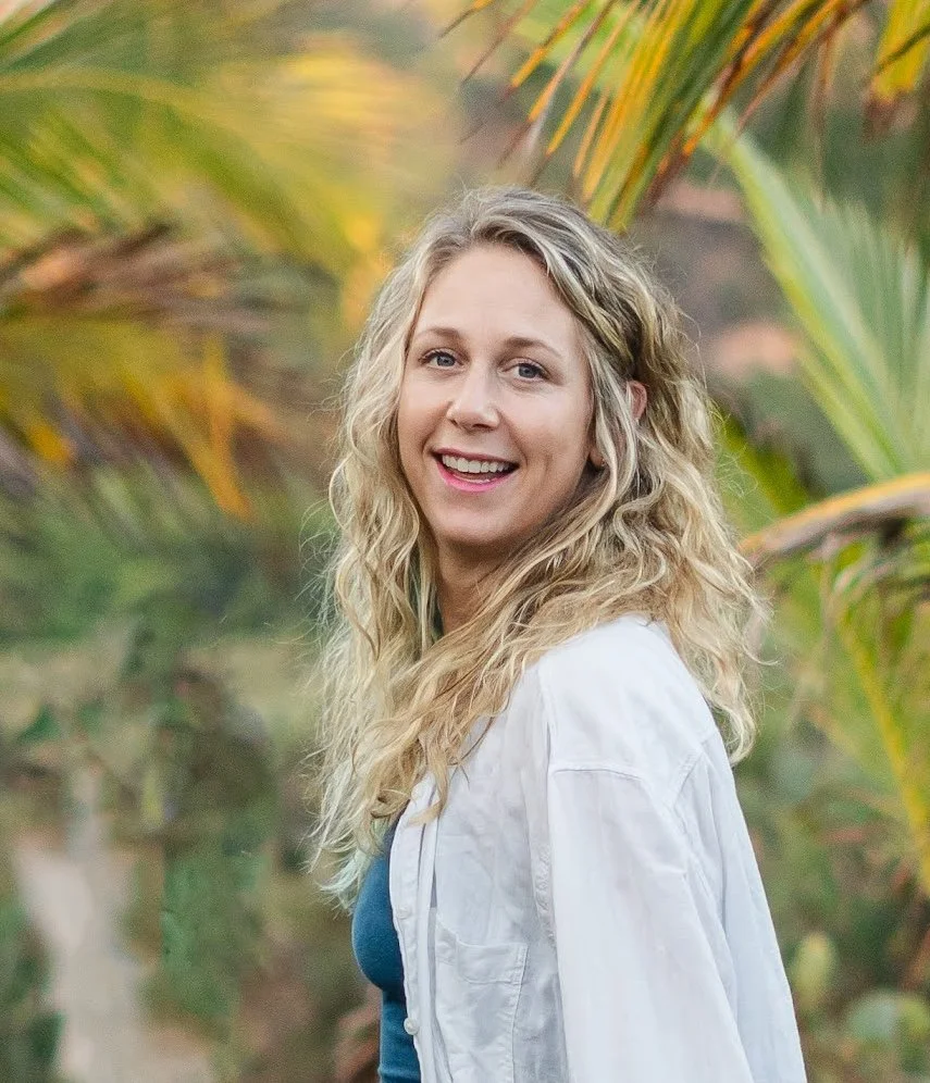 A smiling woman with long, curly blonde hair wearing a white shirt outdoors surrounded by lush green tropical plants.