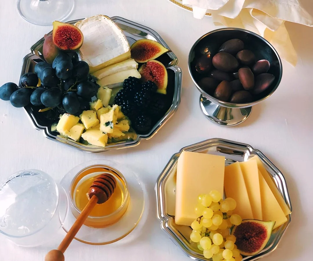 A fruit and cheese platter with grapes, figs, cheese, and honey on a white table, accompanied by bowls of dark grapes and cheese slices, and a jar of honey with a honey dipper.