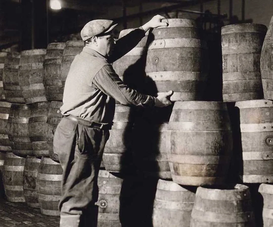 A man in vintage work clothes stacking wooden barrels in a warehouse.
