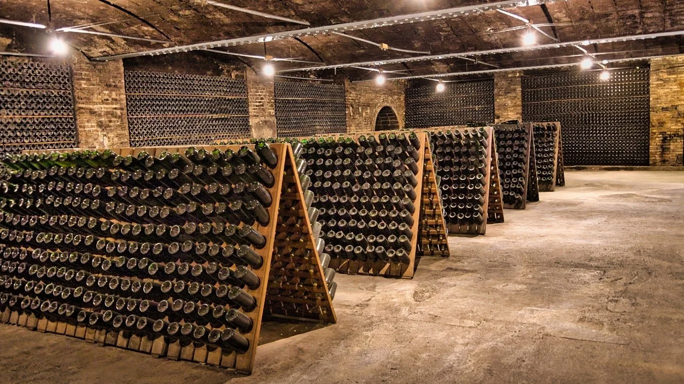 A wine cellar with brick walls and a vaulted ceiling, featuring multiple wooden racks filled with wine bottles.