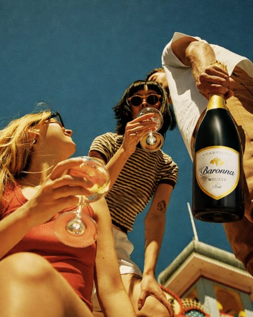 Two women and a man celebrating outdoors under a clear sky, holding glasses of champagne. A large bottle of champagne is being poured. The women are wearing sunglasses, and one has striped shirt. The scene suggests a festive occasion.