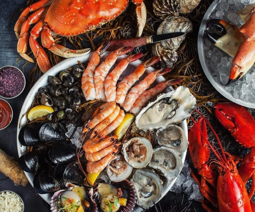 Seafood platter including crab, shrimp, oysters, clams, mussels, lobster, and cocktail sauce, served on ice.
