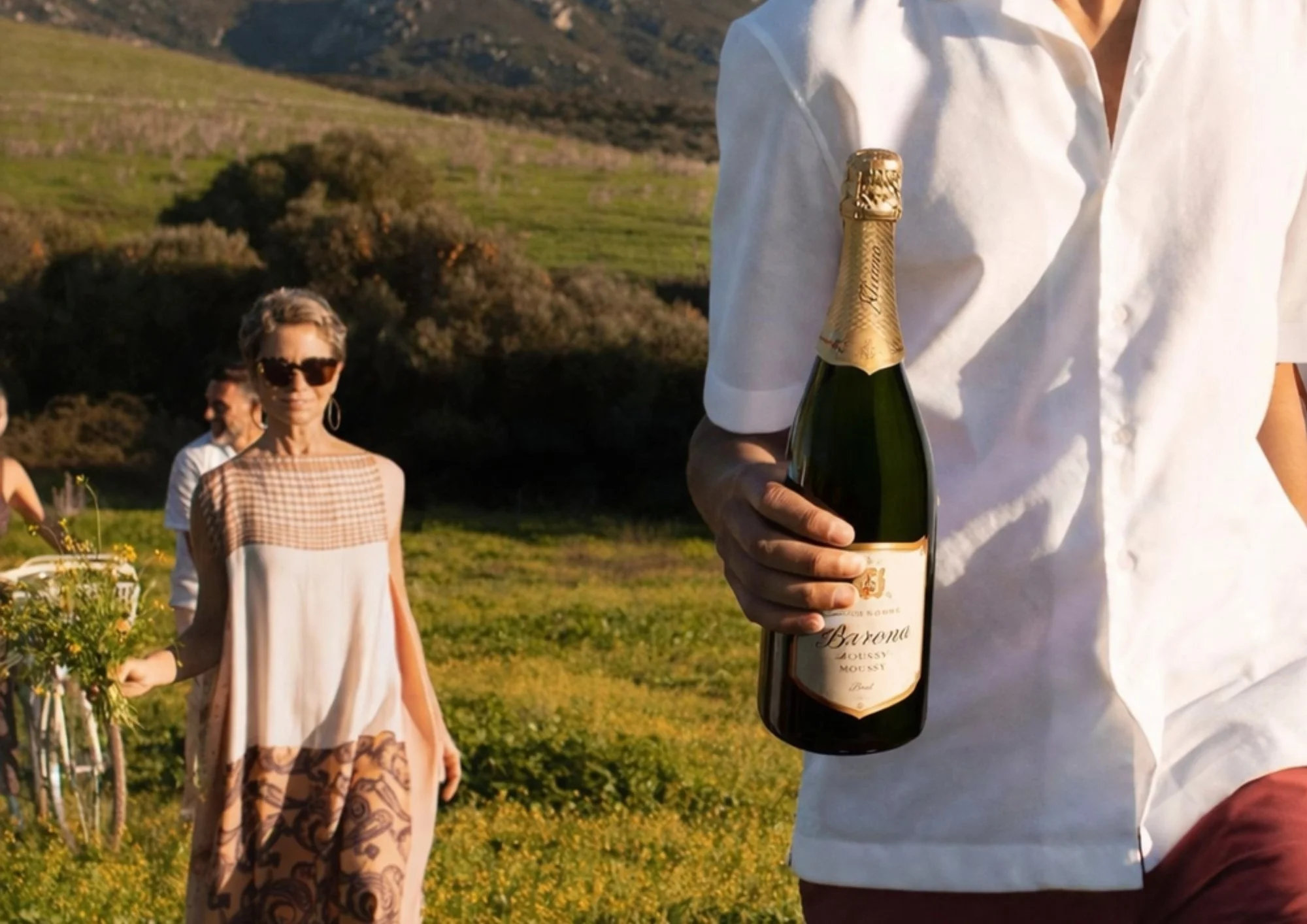 A person in a white shirt holding a bottle of sparkling wine or champagne in an outdoor setting with green hills and scenic landscape. In the background, a woman with short hair, sunglasses, a patterned dress, and earrings stands amidst a field of yellow flowers, with other people nearby.