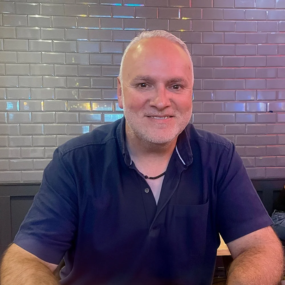 A middle-aged man with short, white hair and a beard, wearing a navy blue shirt, sitting in front of a brick wall with multicolored lighting reflections.