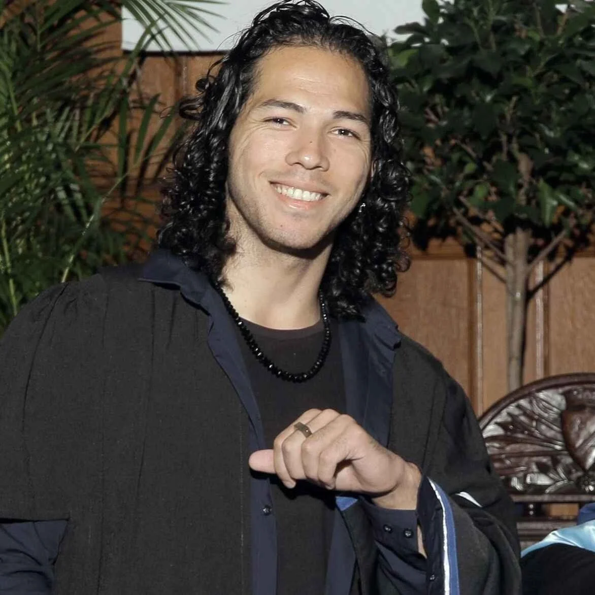 Young man with curly black hair smiling, wearing a black jacket and a beaded necklace, in an outdoor setting with green plants and a wooden fence.