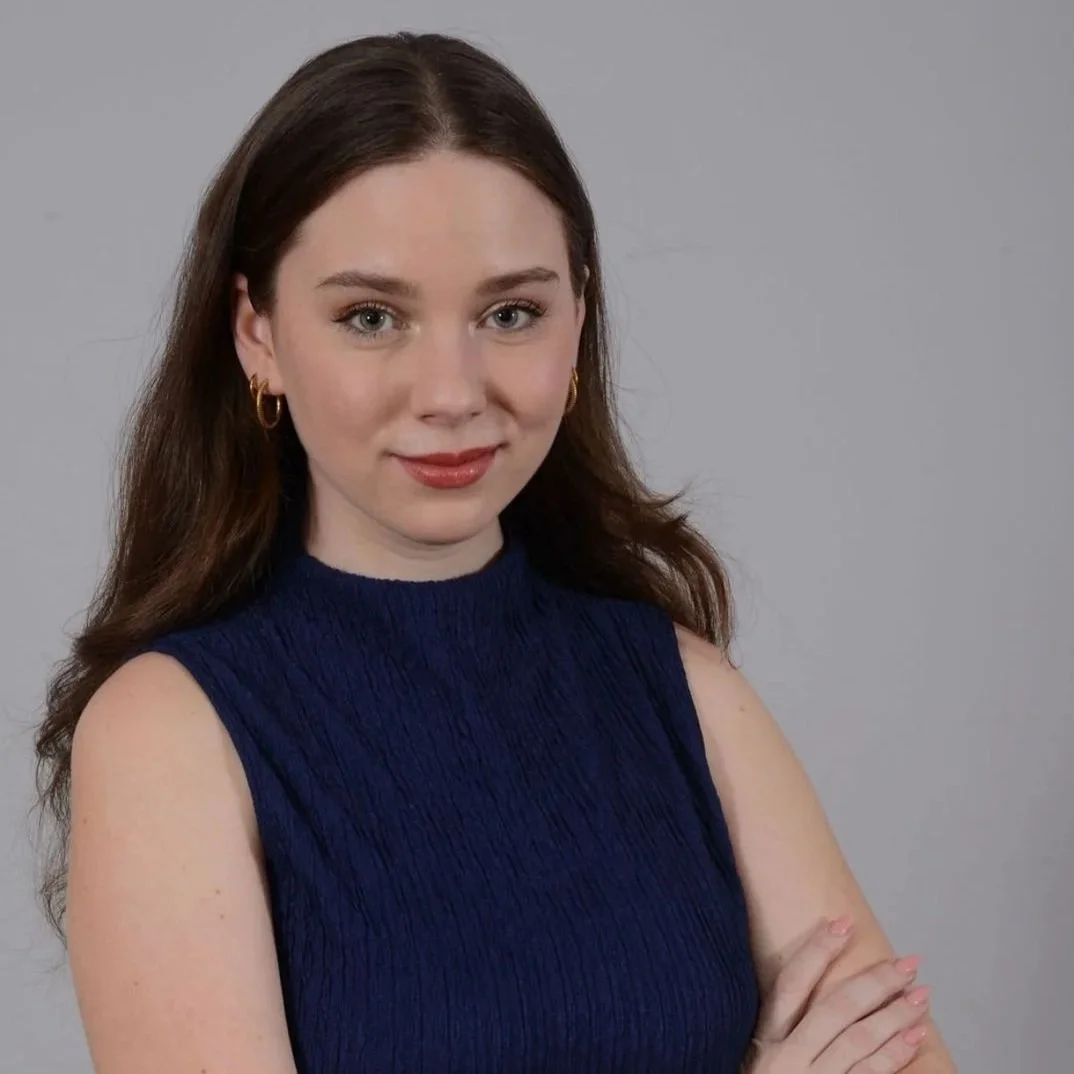 A woman with long brown hair, wearing a sleeveless navy blue top, gold hoop earrings, posing with her arms crossed against a plain gray background.