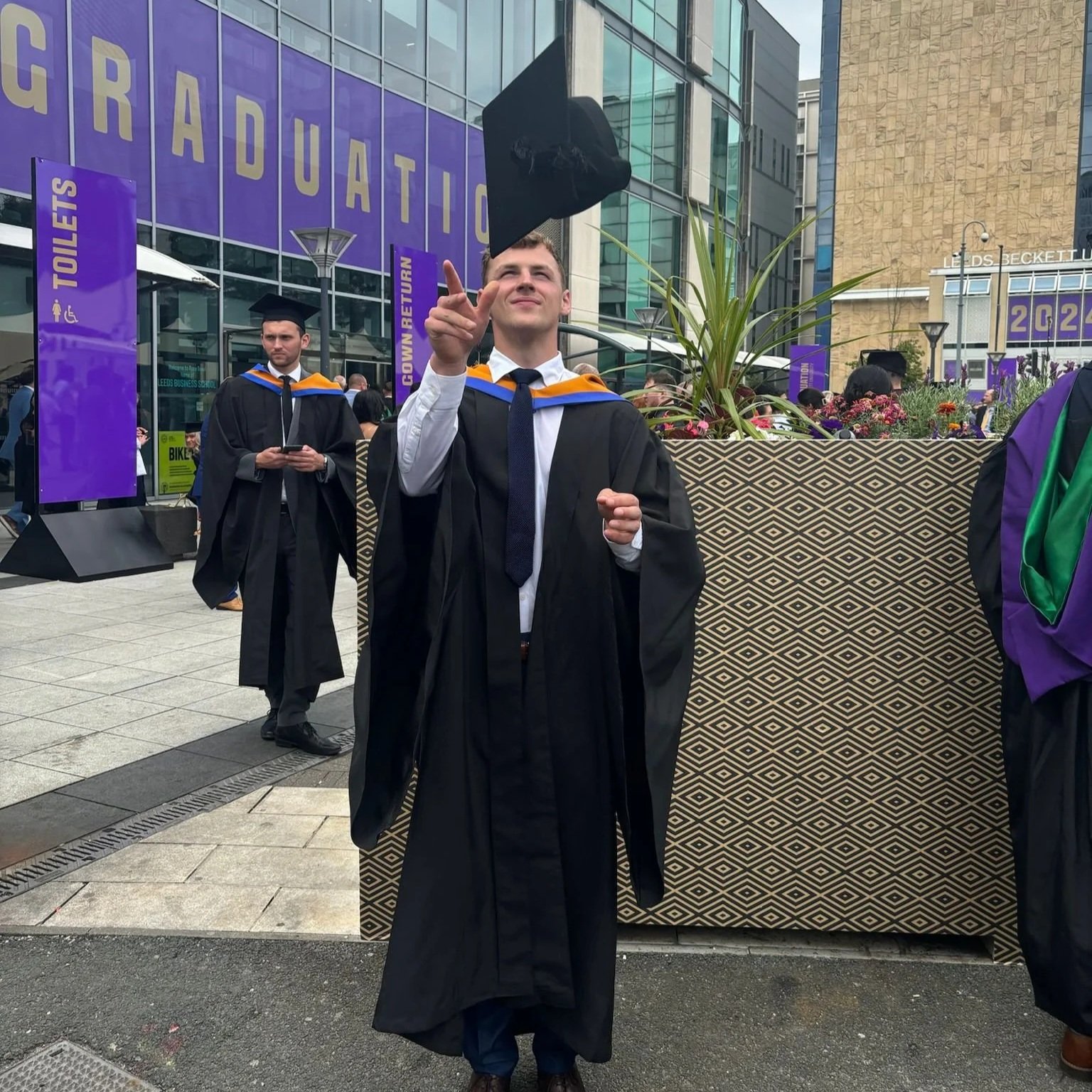 A young man in graduation robes and cap tossing his cap into the air during a graduation ceremony outside a building. Other graduates are visible in the background, and there are signs indicating amenities such as toilets. The scene is lively with people gathered around.