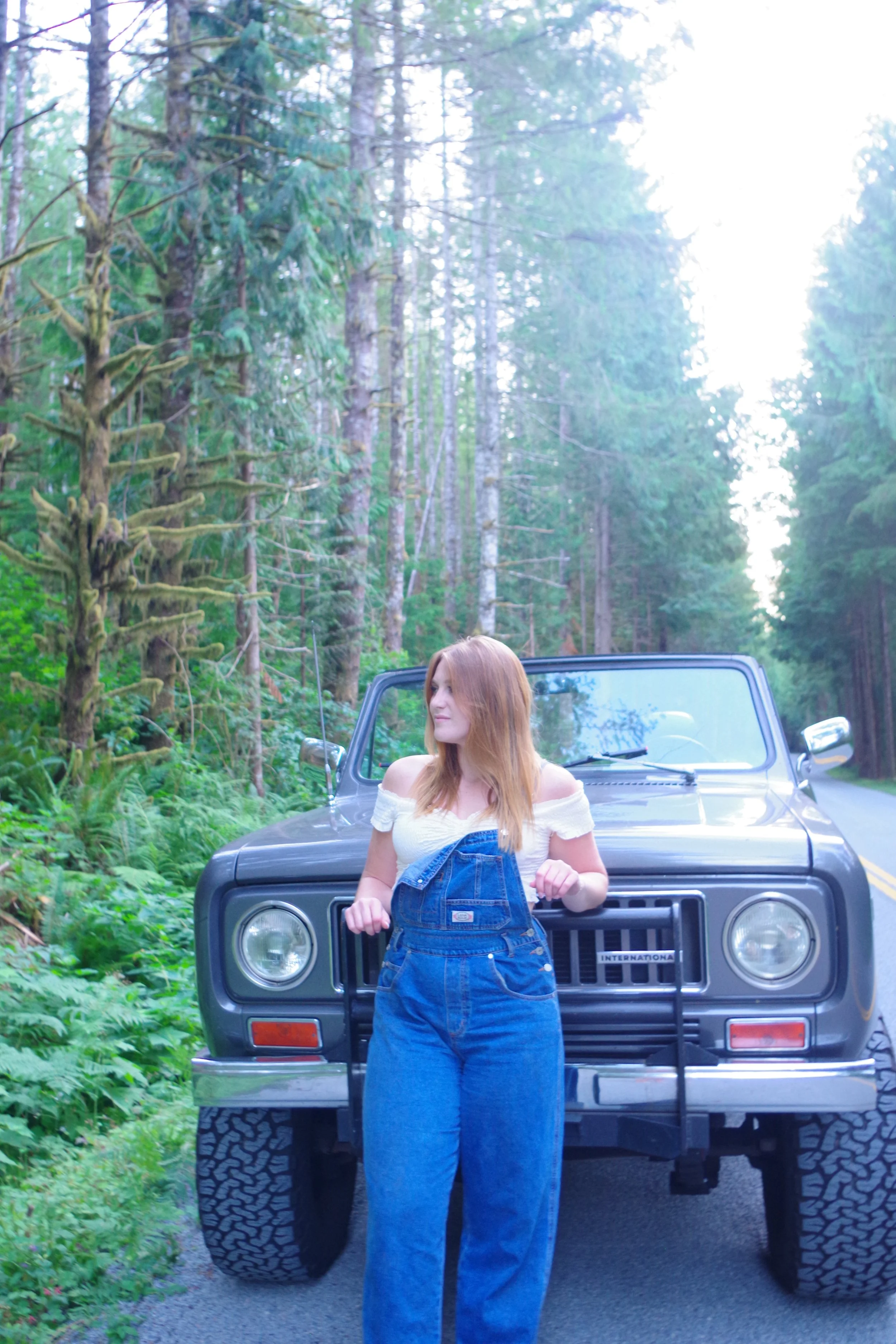 A young woman with red hair standing in front of a dark gray Jeep on a forest road, surrounded by tall trees and greenery.