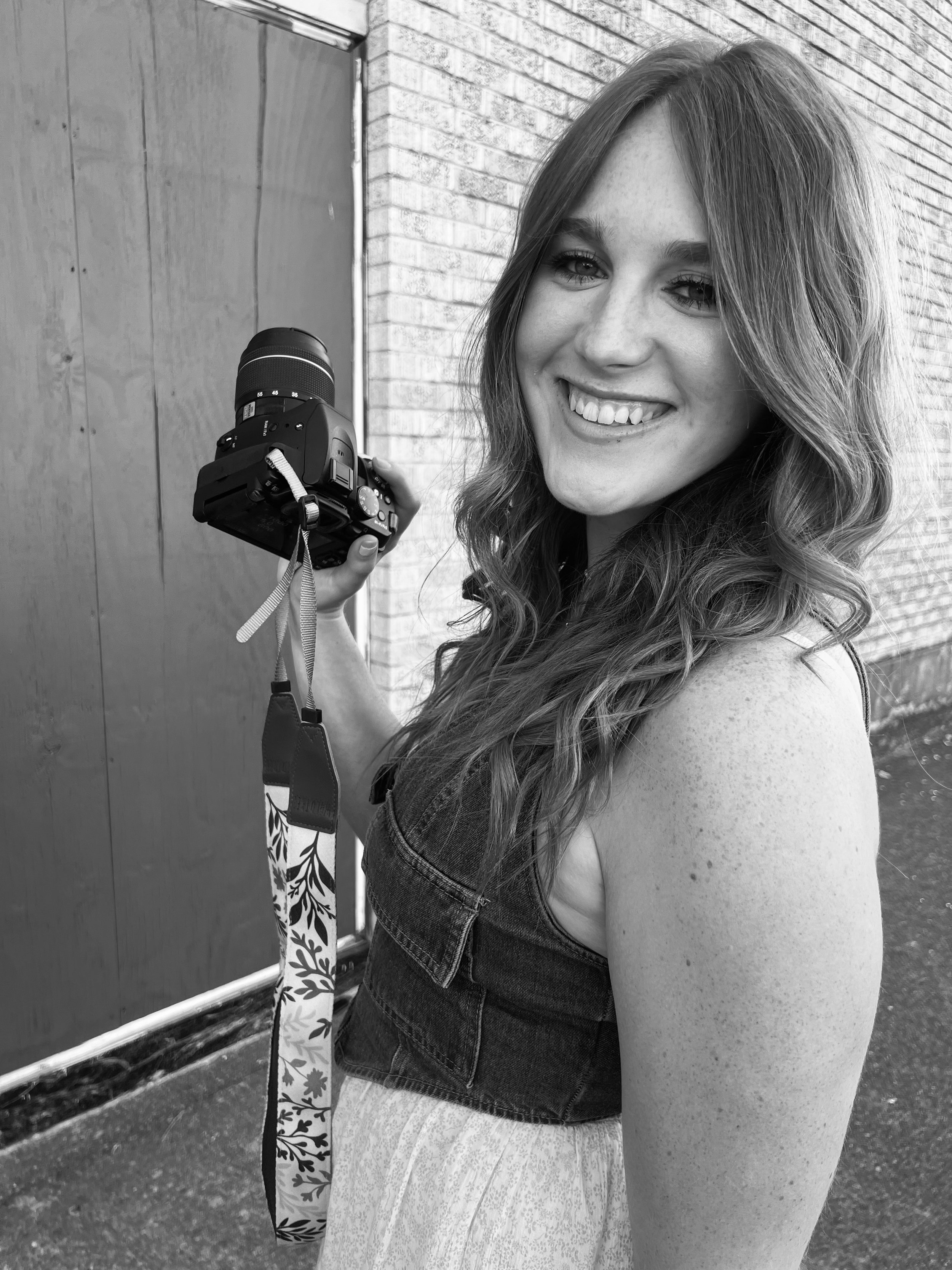 A young woman with wavy hair smiling and holding a camera, standing outdoors near a brick wall.