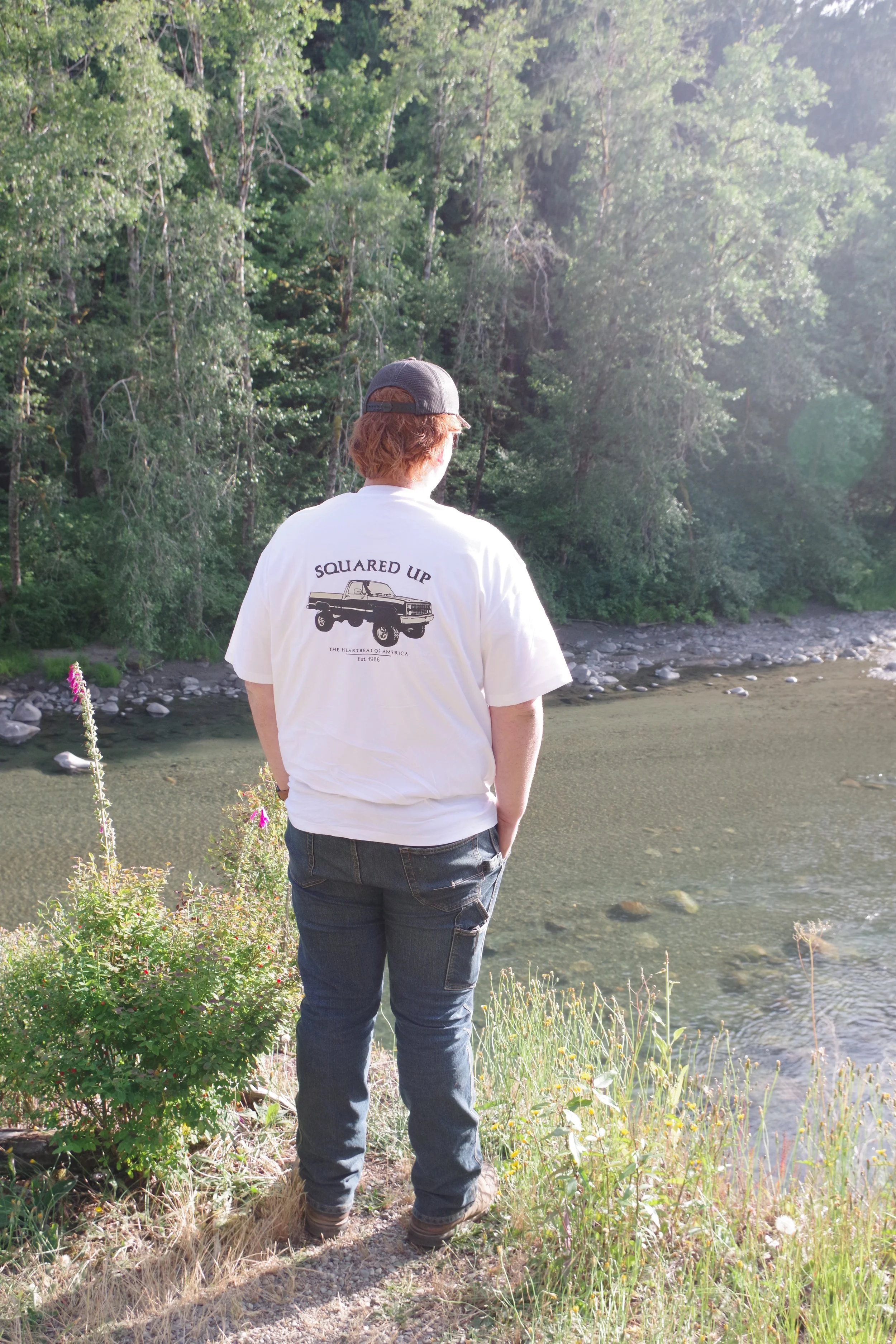 A person with red hair, wearing a black cap, white t-shirt with a graphic of a truck and the words 'SQUARED UP,' and jeans, stands with hands in pockets on a grassy riverbank, looking at the water and forested landscape.
