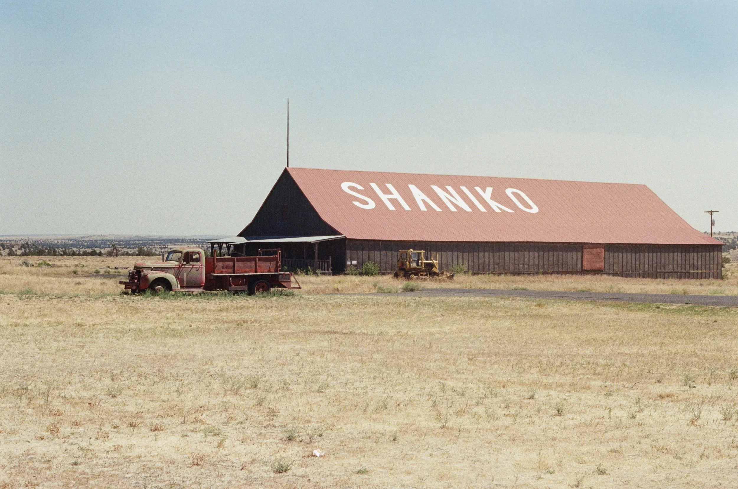 A large barn with the word 'SHANKO' written on its roof, situated in an open, dry field with vintage trucks and a tractor nearby.