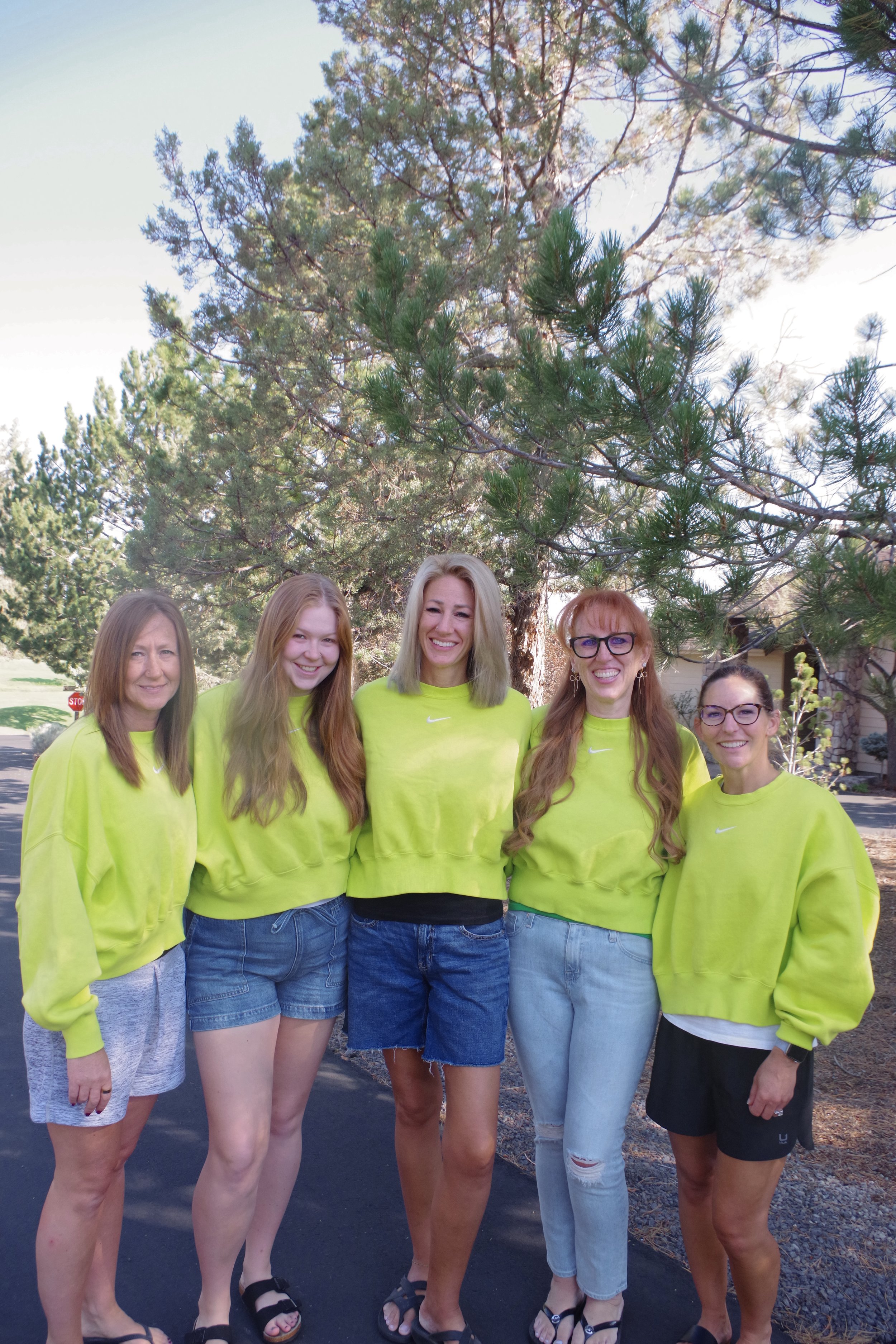 Group of six women standing outdoors in front of a large pine tree, all wearing matching bright yellow sweatshirts and casual shorts or jeans, smiling for the photo.