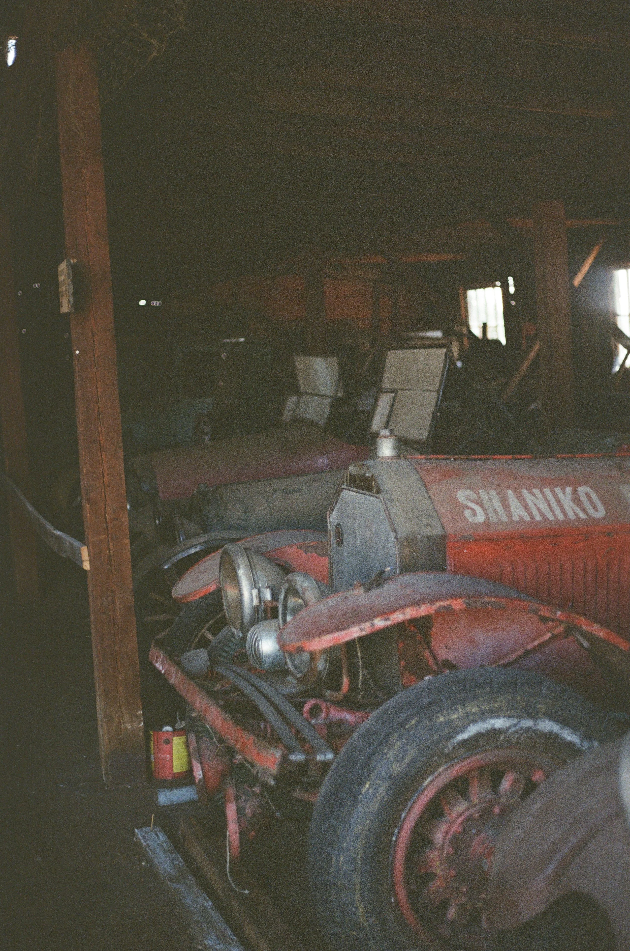 An old red tractor parked inside a barn or shed with wooden beams and walls, a fire extinguisher on the ground near the tractor, and windows letting in some light.
