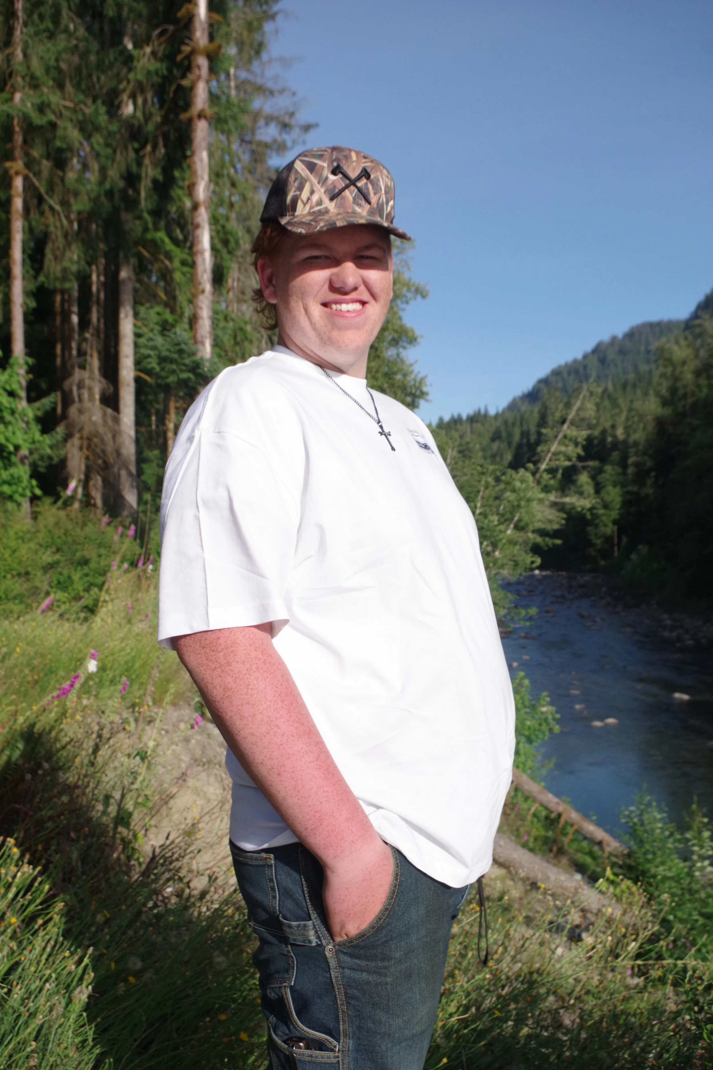 A young man with light skin, freckles, and light brown hair standing outdoors near a river, smiling at the camera. He is wearing a camouflage hat, a white T-shirt, and jeans, with his hand in his pocket. The background features trees, a river, and mo