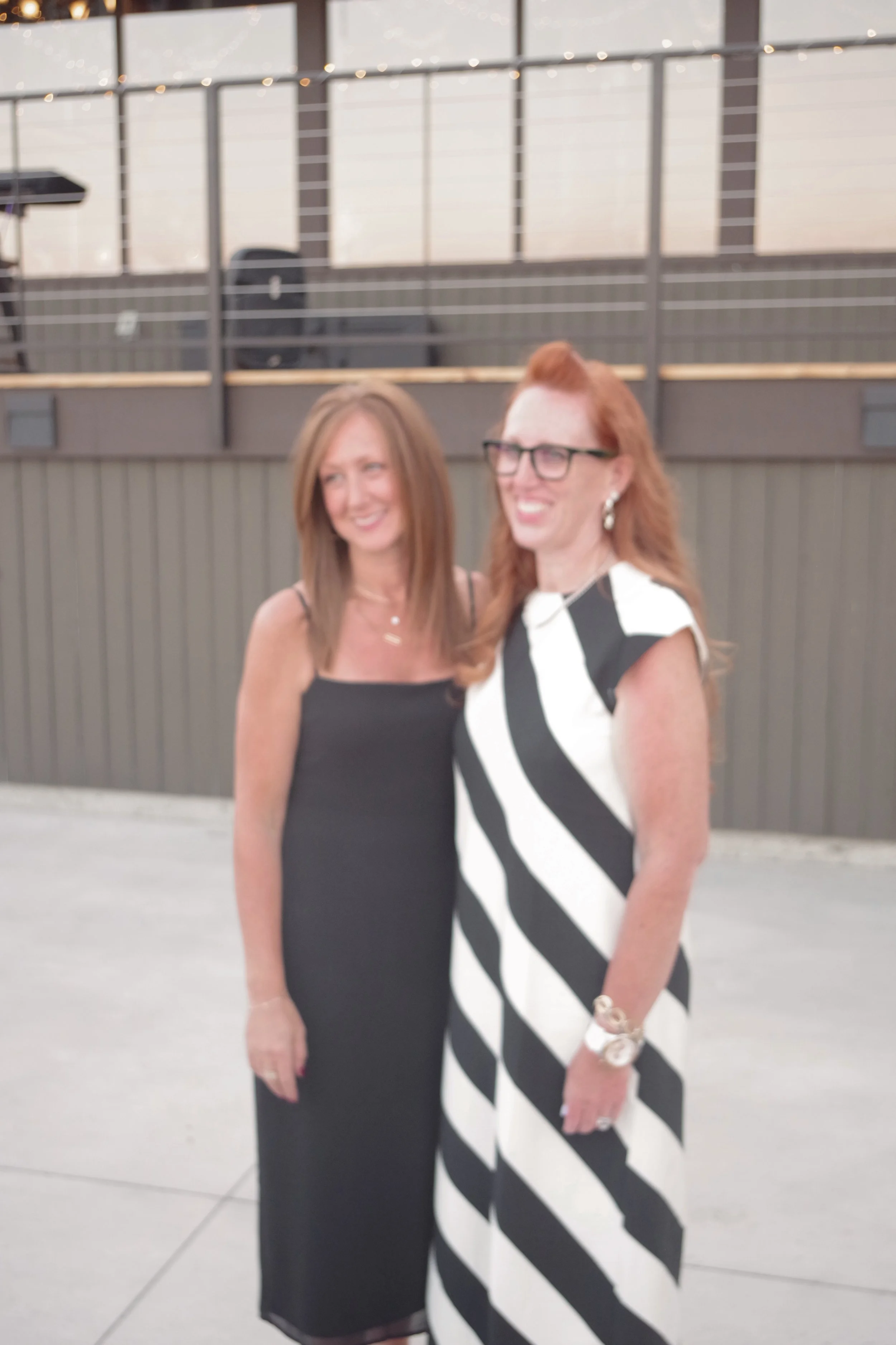 Two women standing together in an outdoor setting, smiling. One woman wears a black dress, the other wears a black-and-white striped dress. The background features a building with large windows and metal railings.