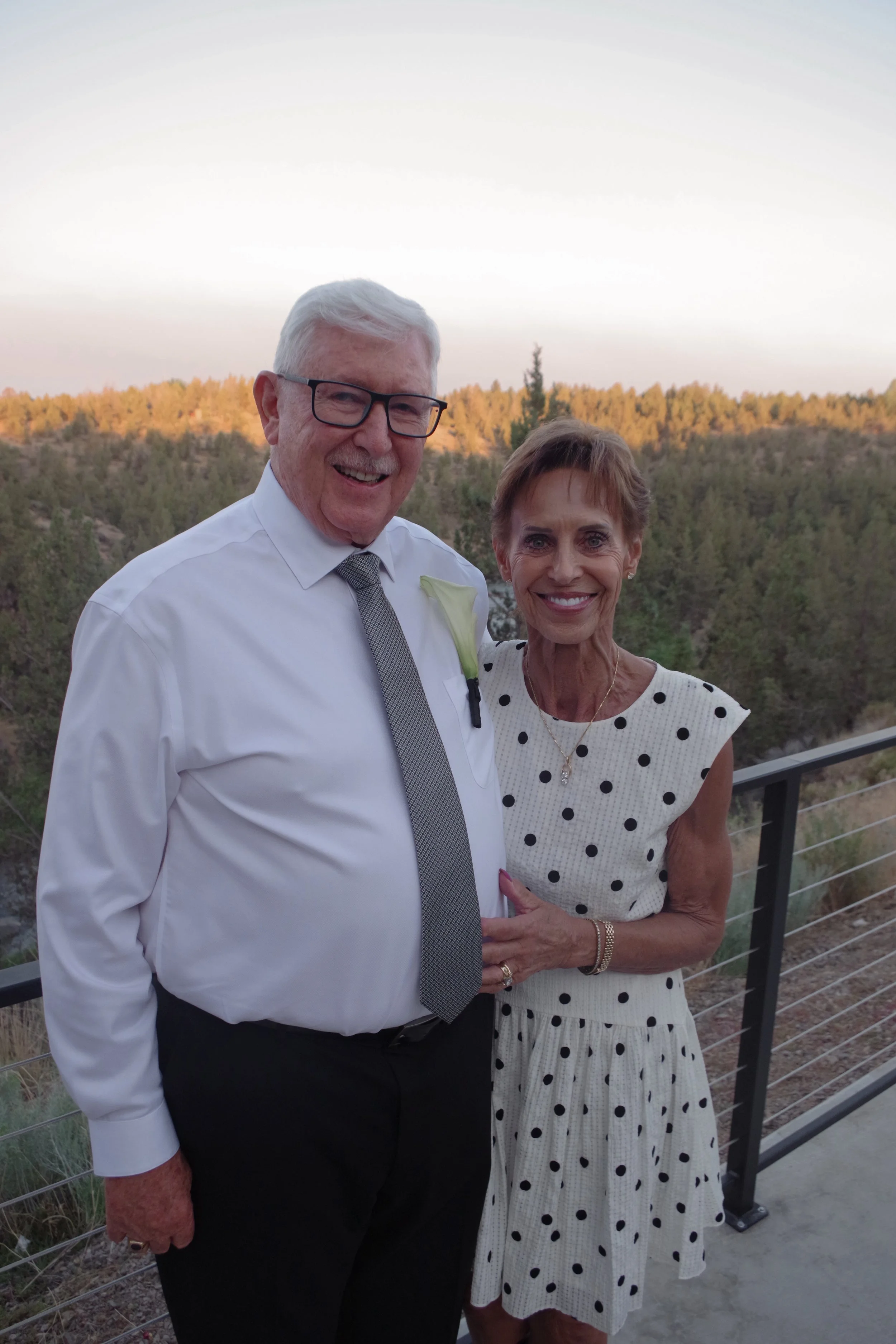 A smiling elderly man with white hair, glasses, a white shirt, a gray tie, and a white boutonnière standing next to a smiling elderly woman with short brown hair, wearing a white dress with black polka dots, on a balcony with a mountain landscape in 