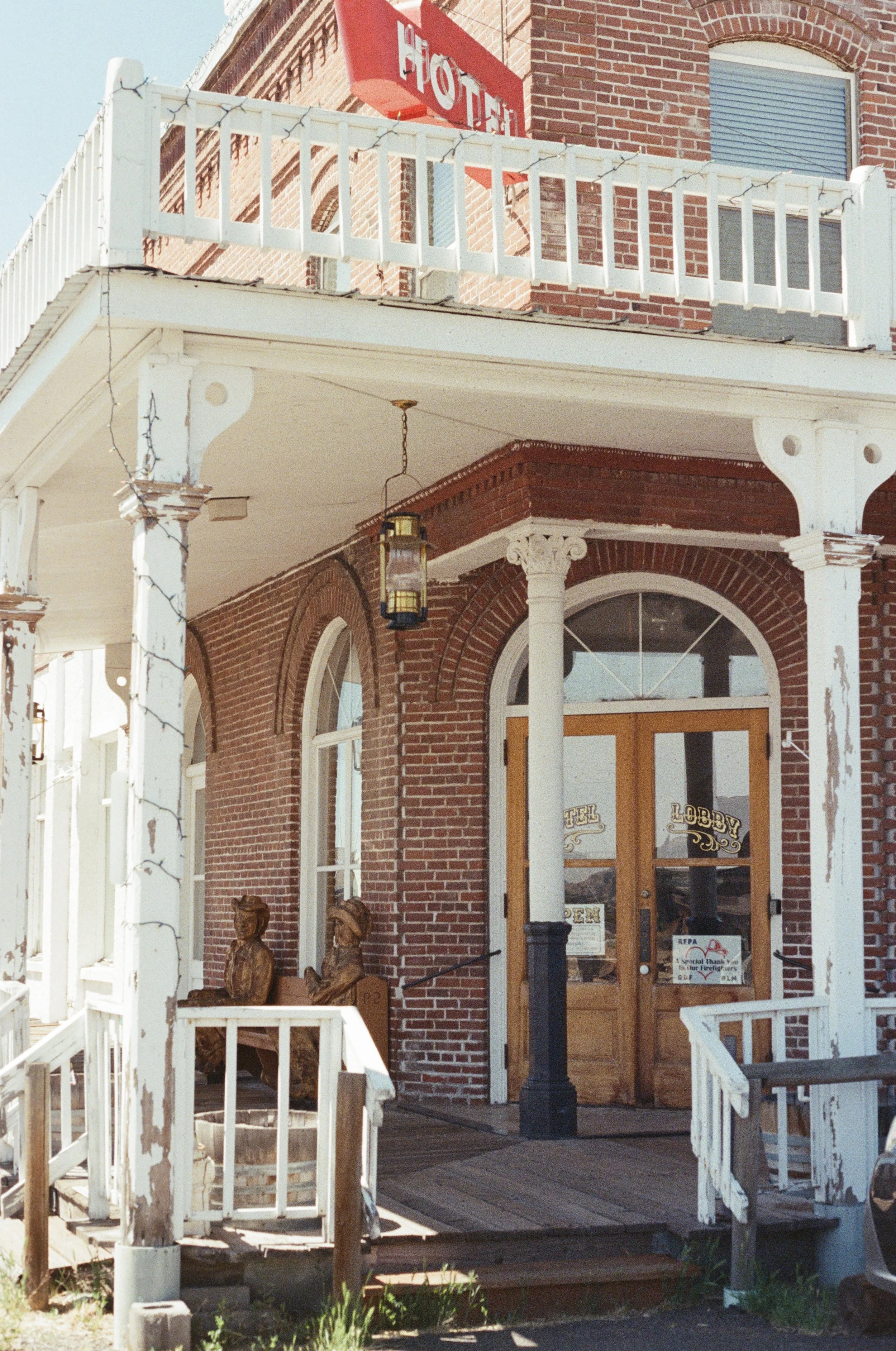 Red brick building with a balcony, a sign that reads 'HOTEL' in white letters on a red background, white porch railing with string lights, a hanging lantern, sculptures of animals, and a glass door with signs indicating the hotel is open.