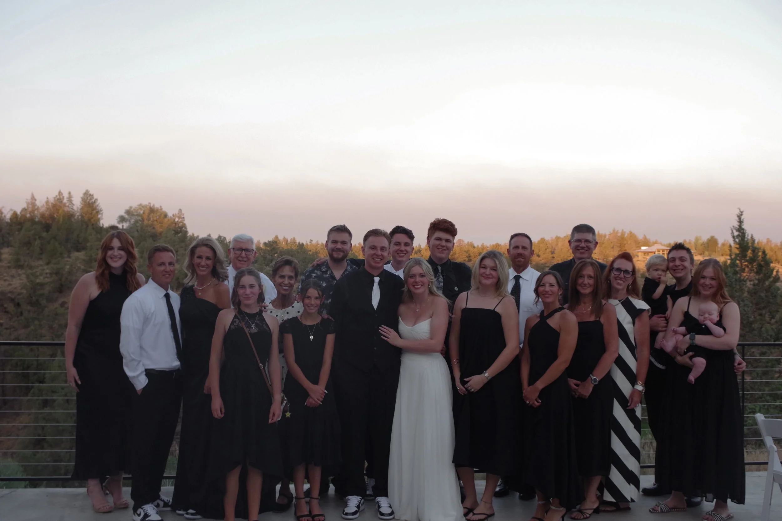 Group of people including adults and children posing outdoors during sunset with trees and a house in the background.