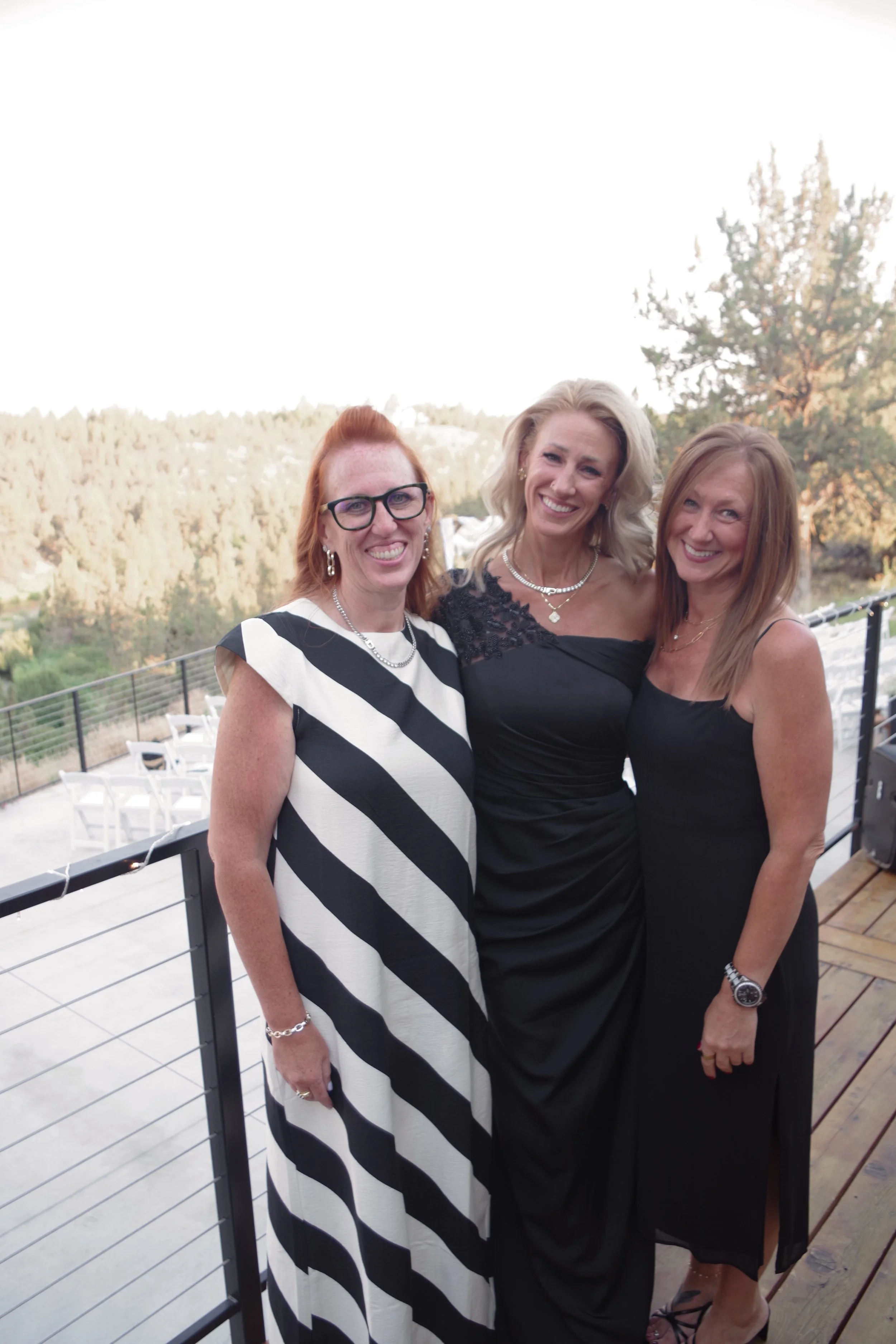 Three women smiling outdoors on a deck with a scenic background of trees and hills, dressed in formal attire.