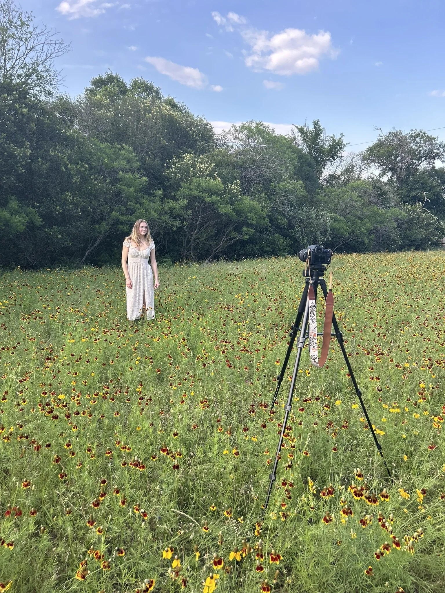 A woman standing in a field of yellow and red flowers with a camera on a tripod in front of her, and a background of trees and a partly cloudy sky.