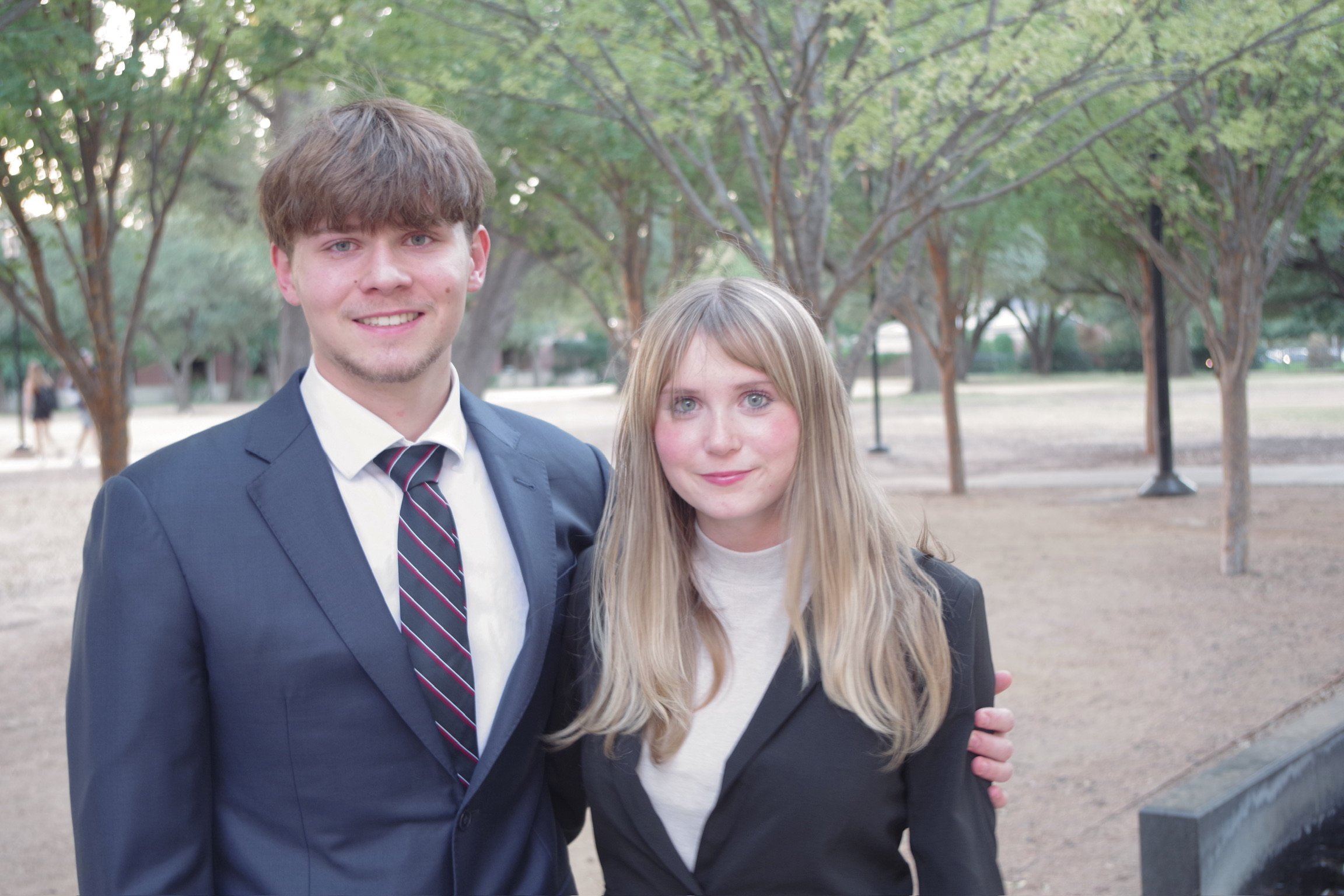 A young man in a suit and tie standing next to a young woman in a blazer, both smiling outdoors among trees.