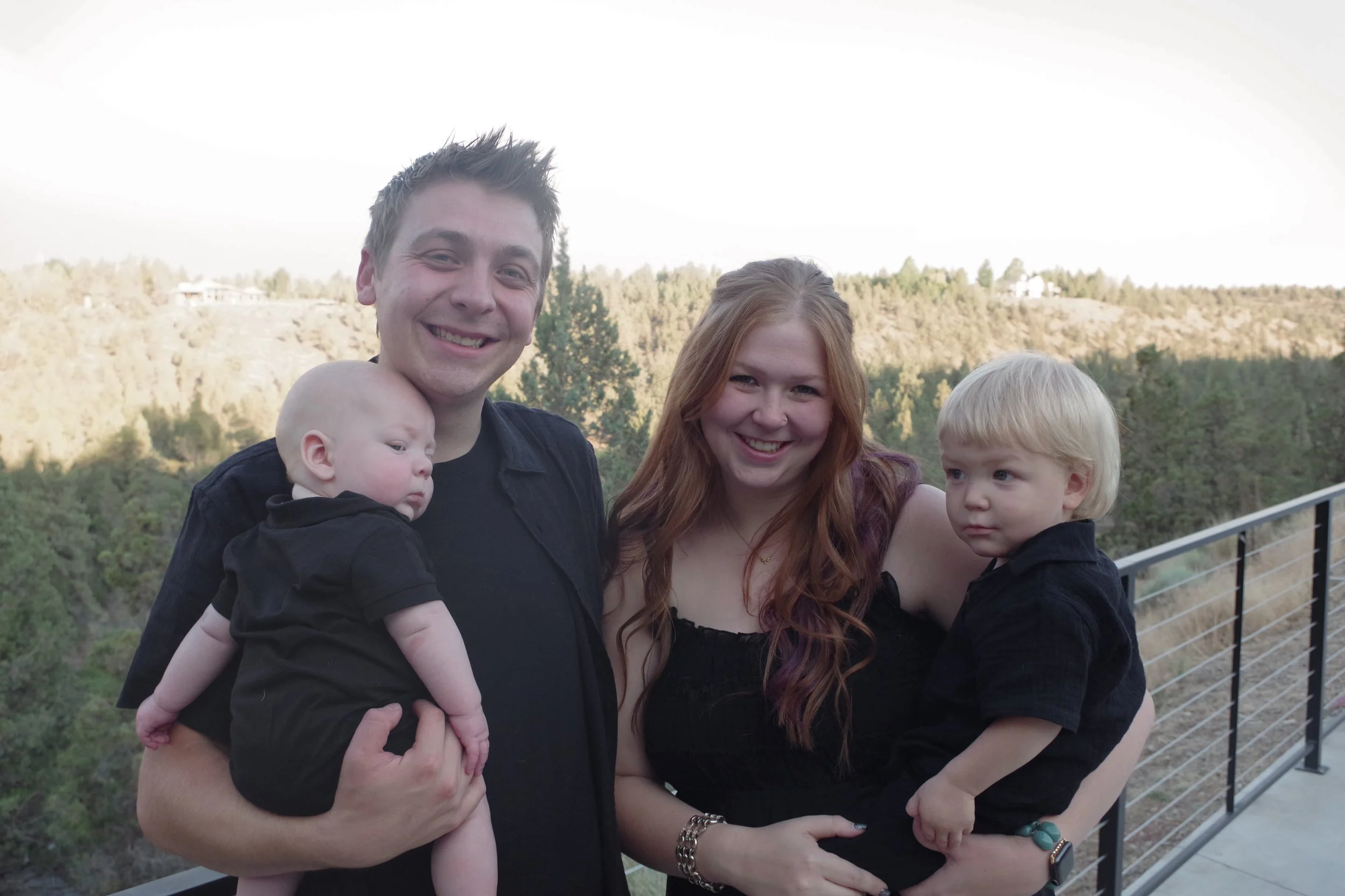 A smiling man holding a baby, a smiling woman, and a young child with blonde hair standing outside on a balcony with a scenic landscape of trees and hills in the background.