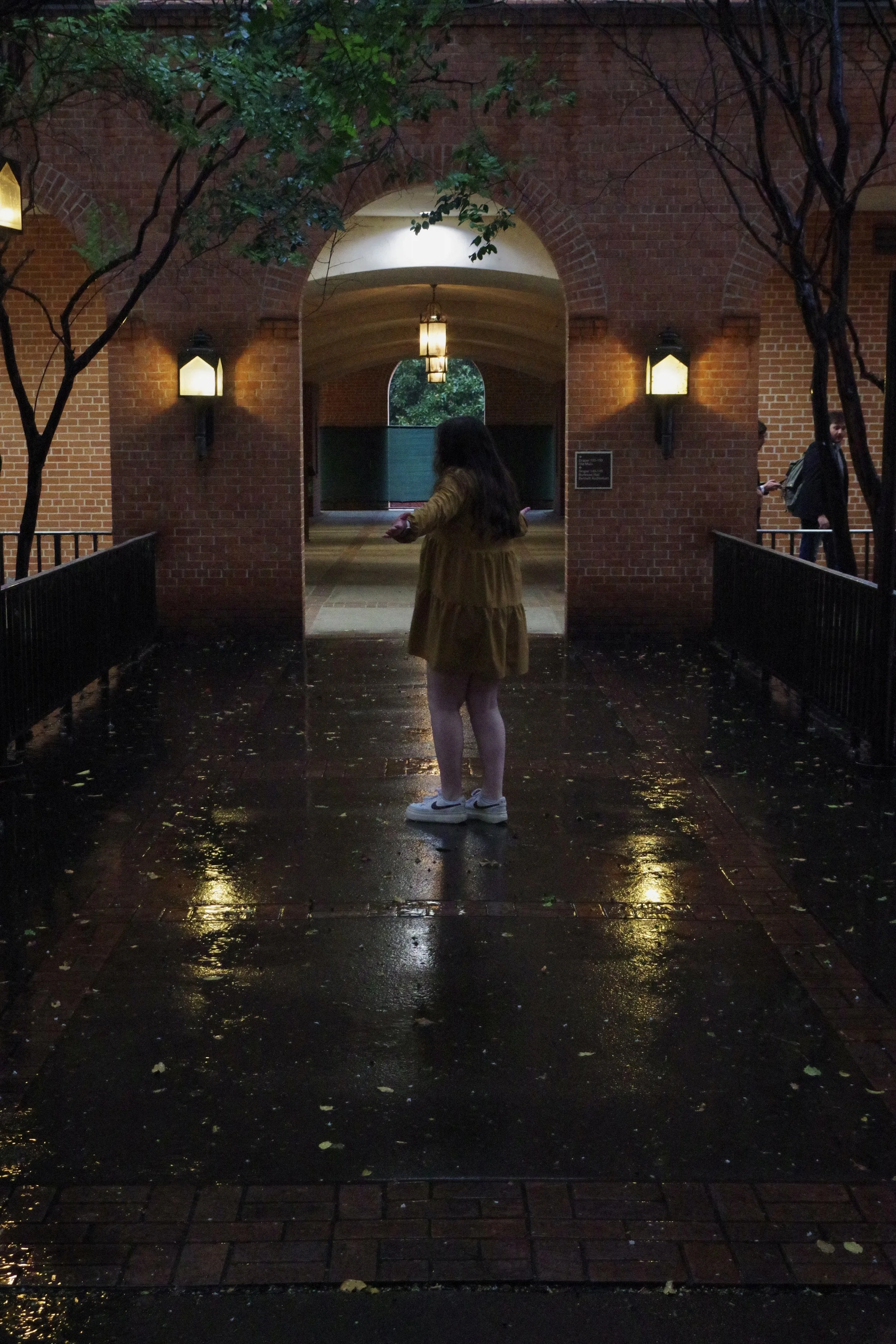 A woman with long dark hair wearing a yellow coat and white sneakers standing on a wet brick sidewalk at night, under an arched brick structure with black lanterns, trees on either side, and a person with a backpack walking in the background.