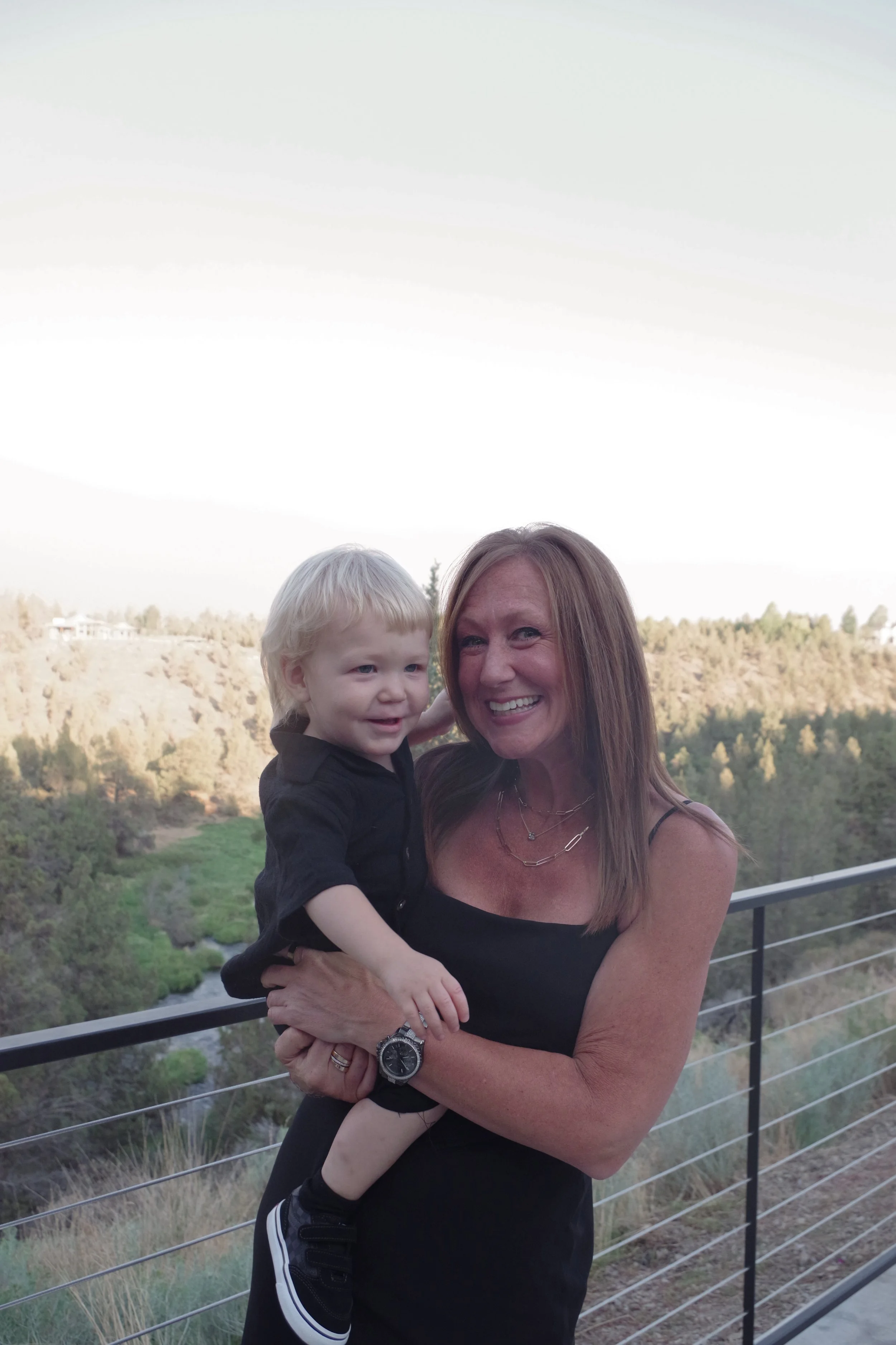 A woman with long, light brown hair and a black sleeveless dress is holding a young blonde-haired boy in her arms. They are standing on a balcony with a metal railing, overlooking a landscape of trees and a river. The woman is smiling, and the boy is