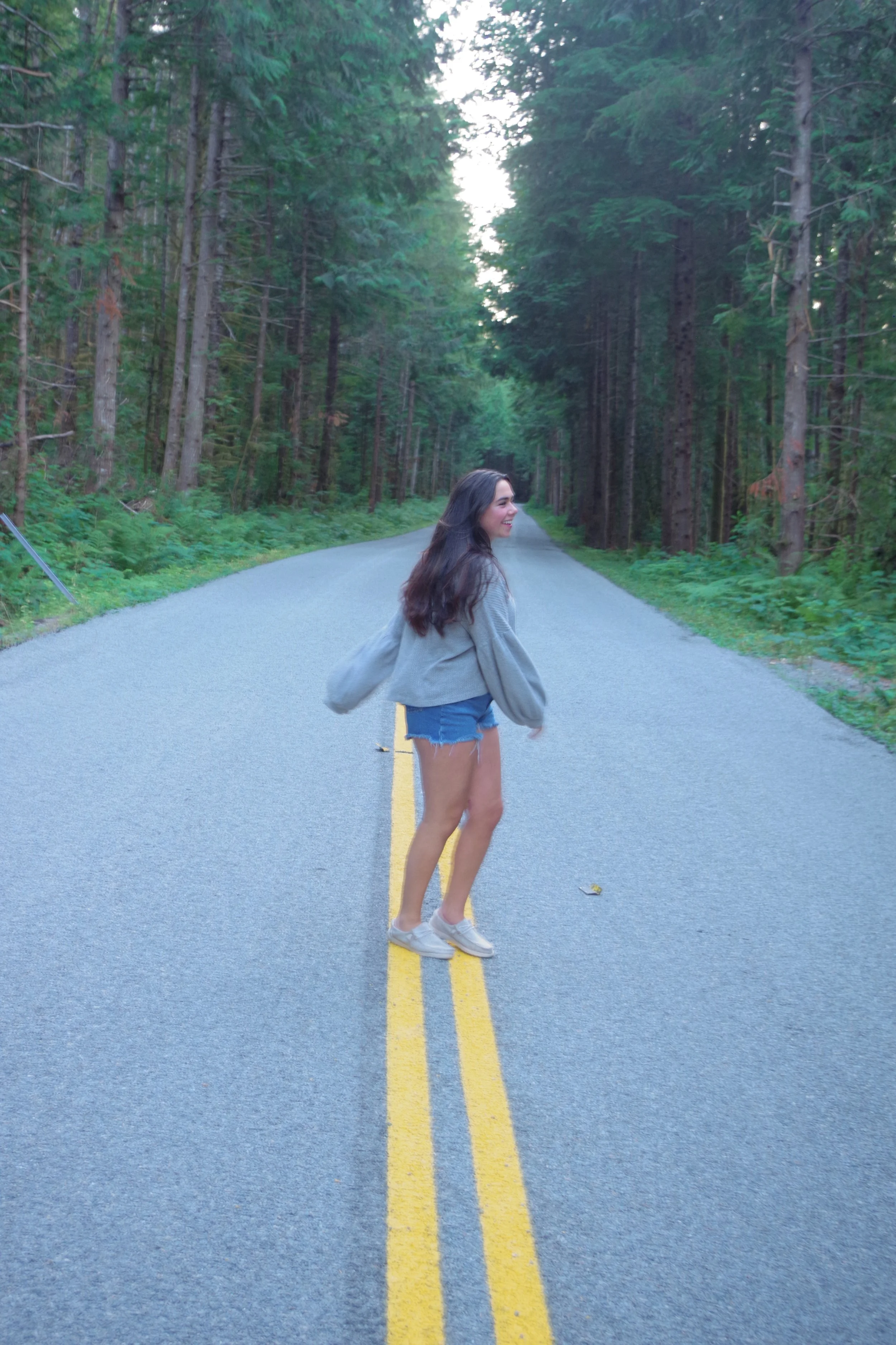 A woman in denim shorts, a gray sweater, and white sneakers standing on a rural road surrounded by tall green trees, smiling and looking to the side.
