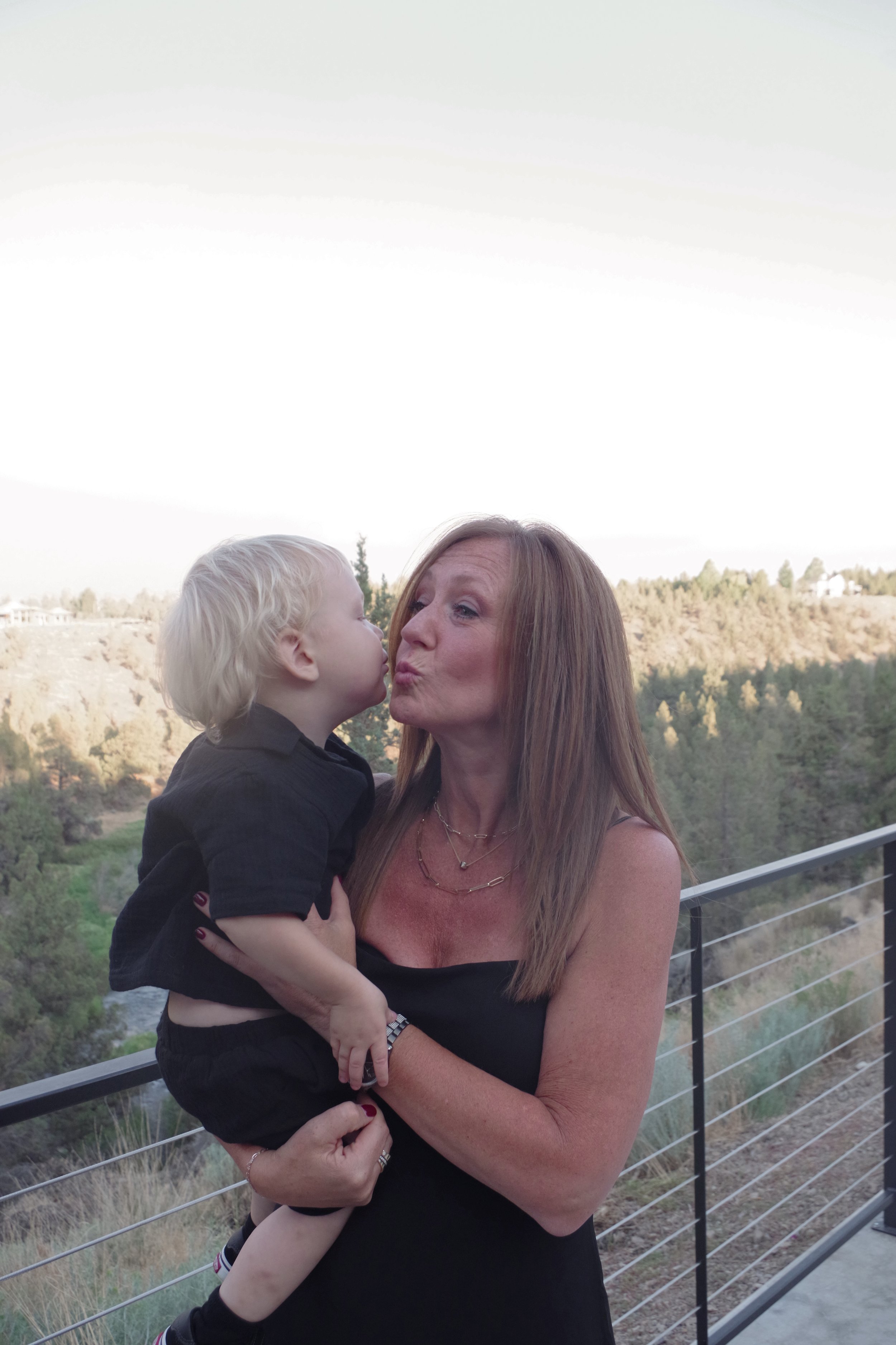 A woman with long red hair holding a young blond boy who is giving her a kiss on the lips outdoors with a scenic background of trees and a cloudy sky.