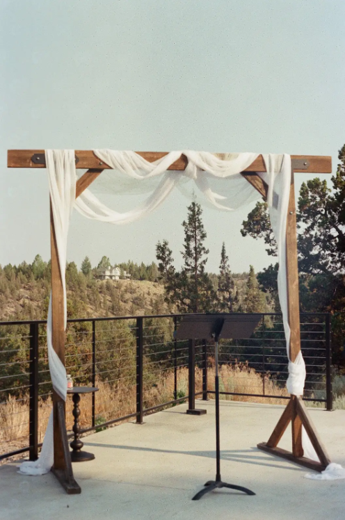 Wooden wedding arch decorated with white fabric, set on a balcony overlooking a forested landscape.