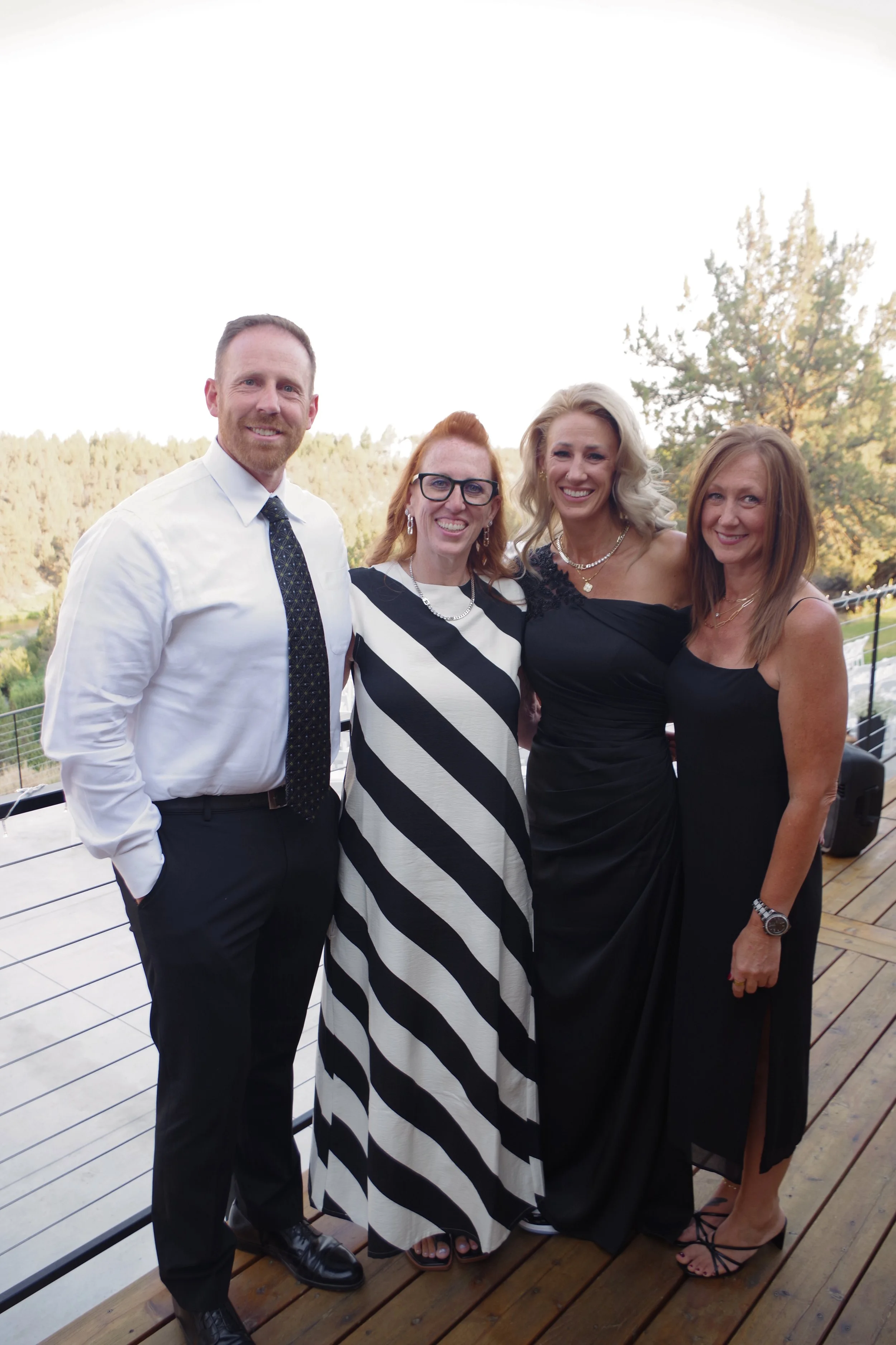 Four people standing outdoors on a wooden deck with a railing, smiling at the camera. The background shows trees and a partly cloudy sky.