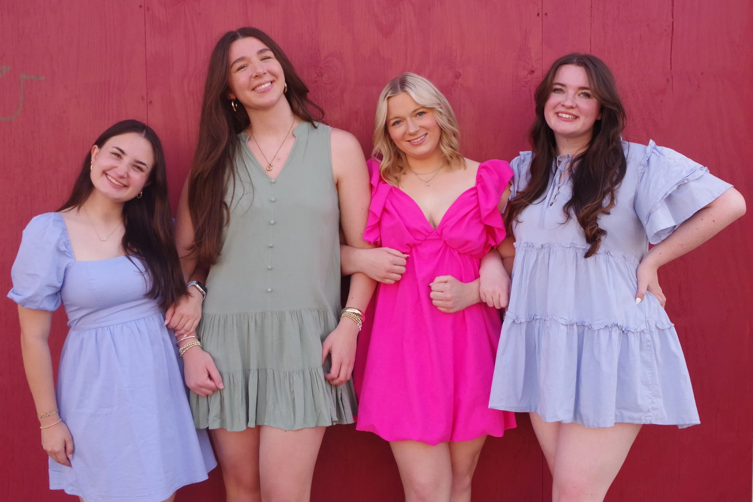 Five women standing arm in arm, smiling, in front of a red wooden wall, wearing colorful dresses in pastel and bright pink hues.