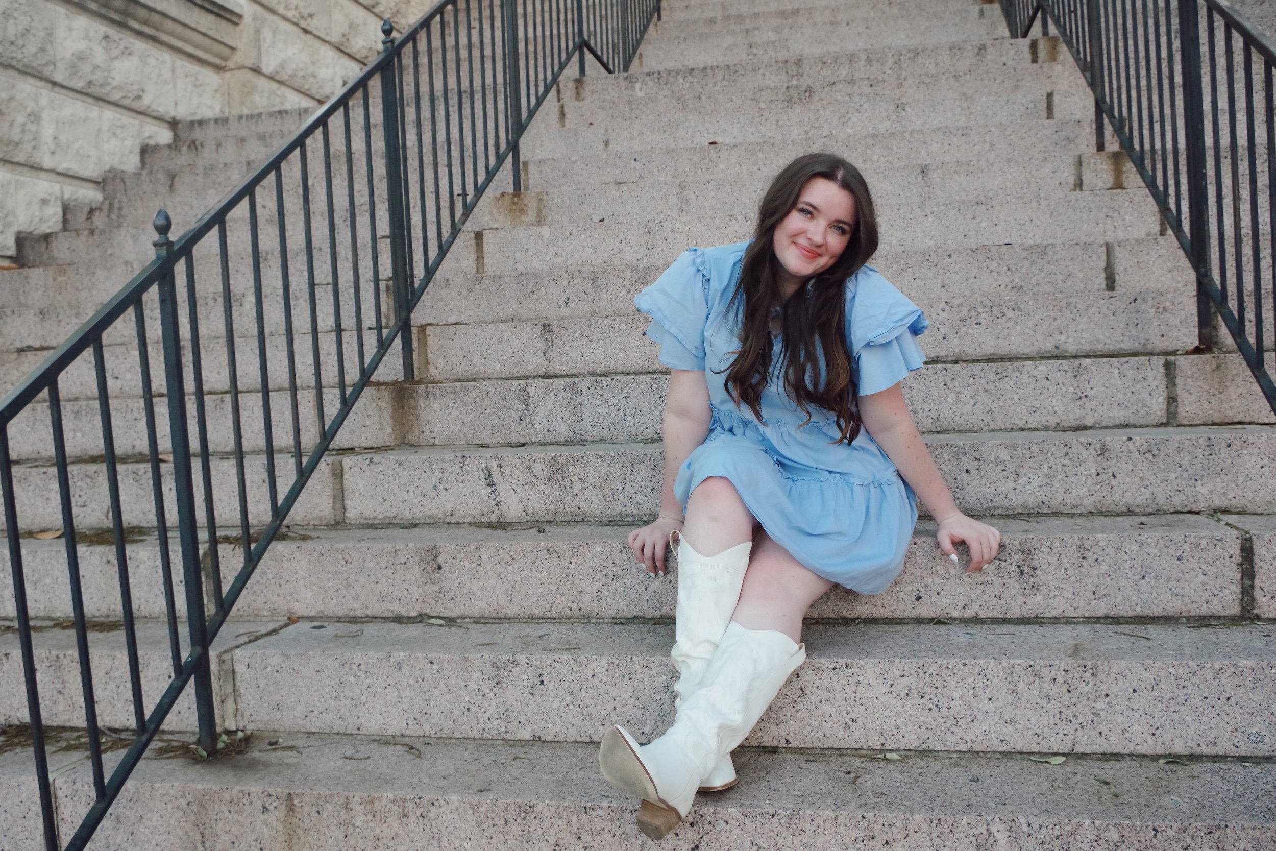 A young woman with long dark hair is sitting on stone steps outdoors. She is smiling and wearing a light blue dress and white cowboy boots.