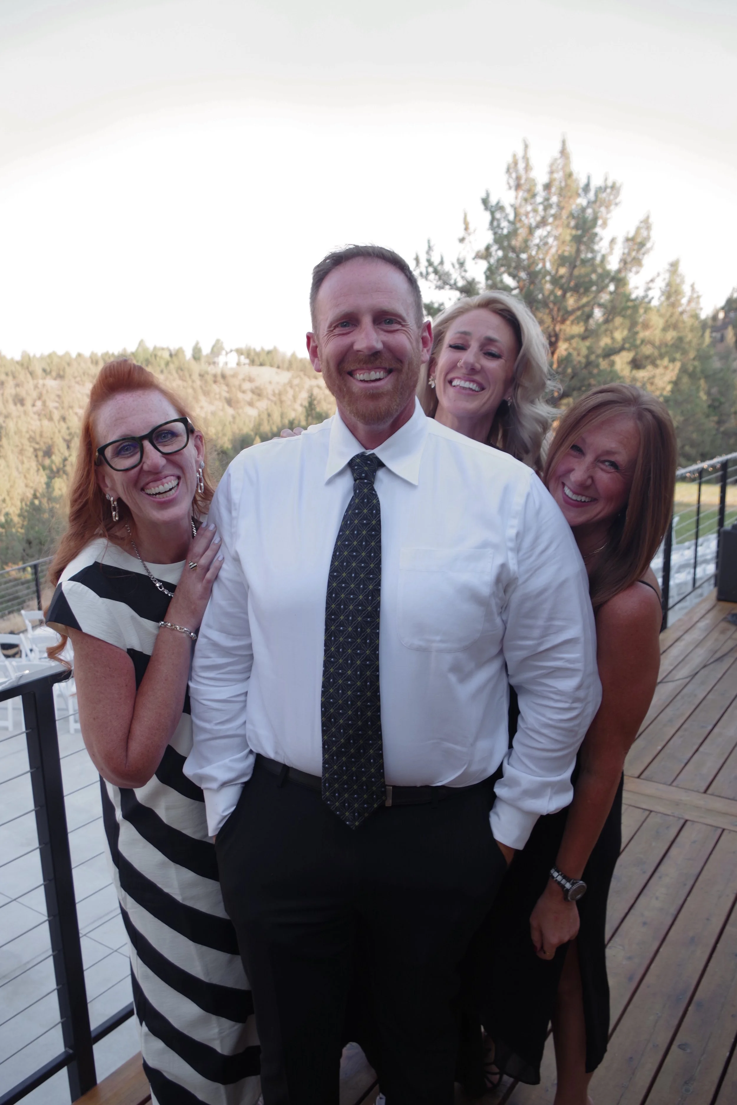 Group of four smiling adults in formal attire standing outdoors on a wooden deck, with trees and hills in the background.