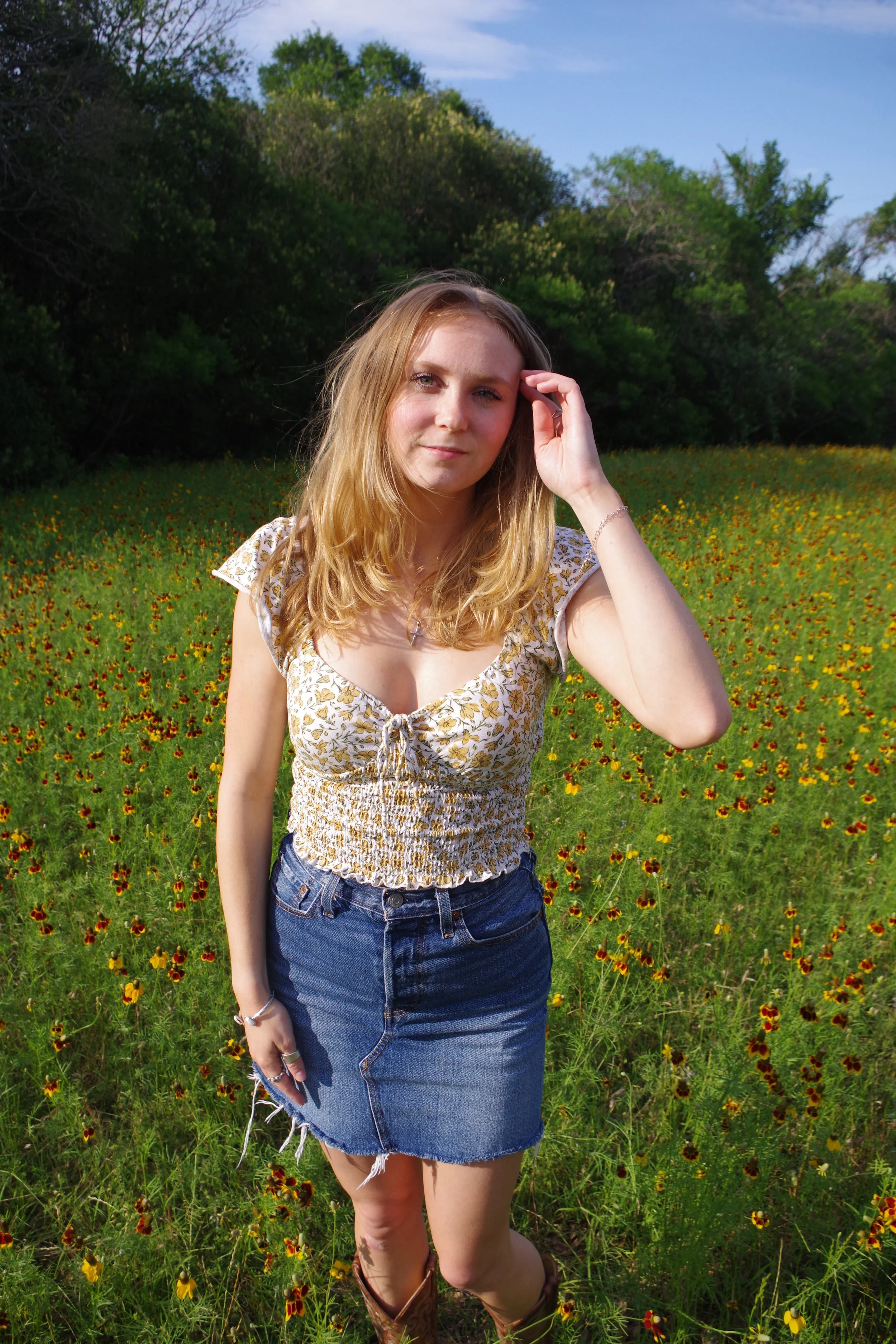A young woman with blonde hair standing in a field of yellow and brown flowers, with green trees and a blue sky in the background.