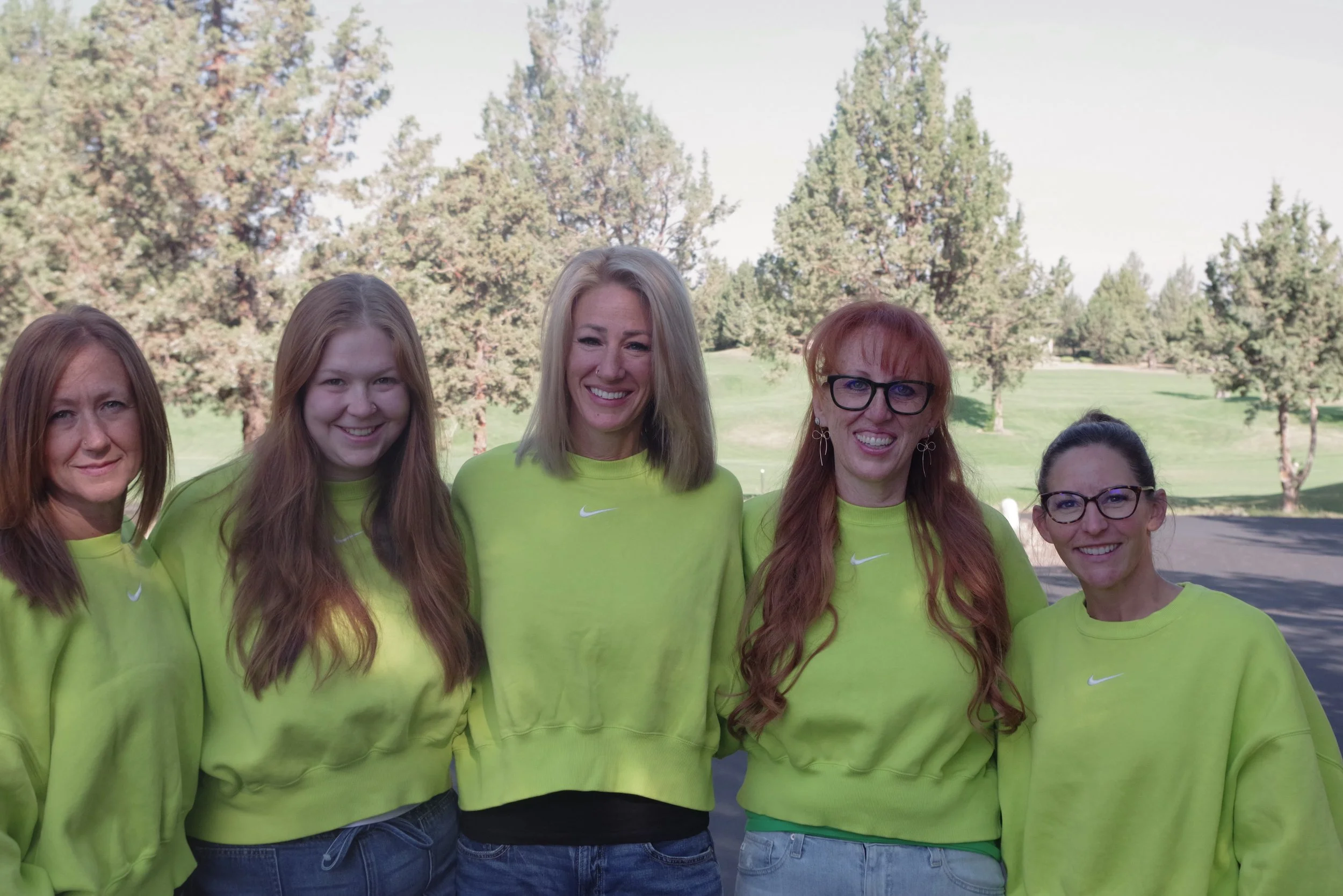 Group of women wearing matching bright yellow Nike shirts, standing outdoors in a park with trees in the background.