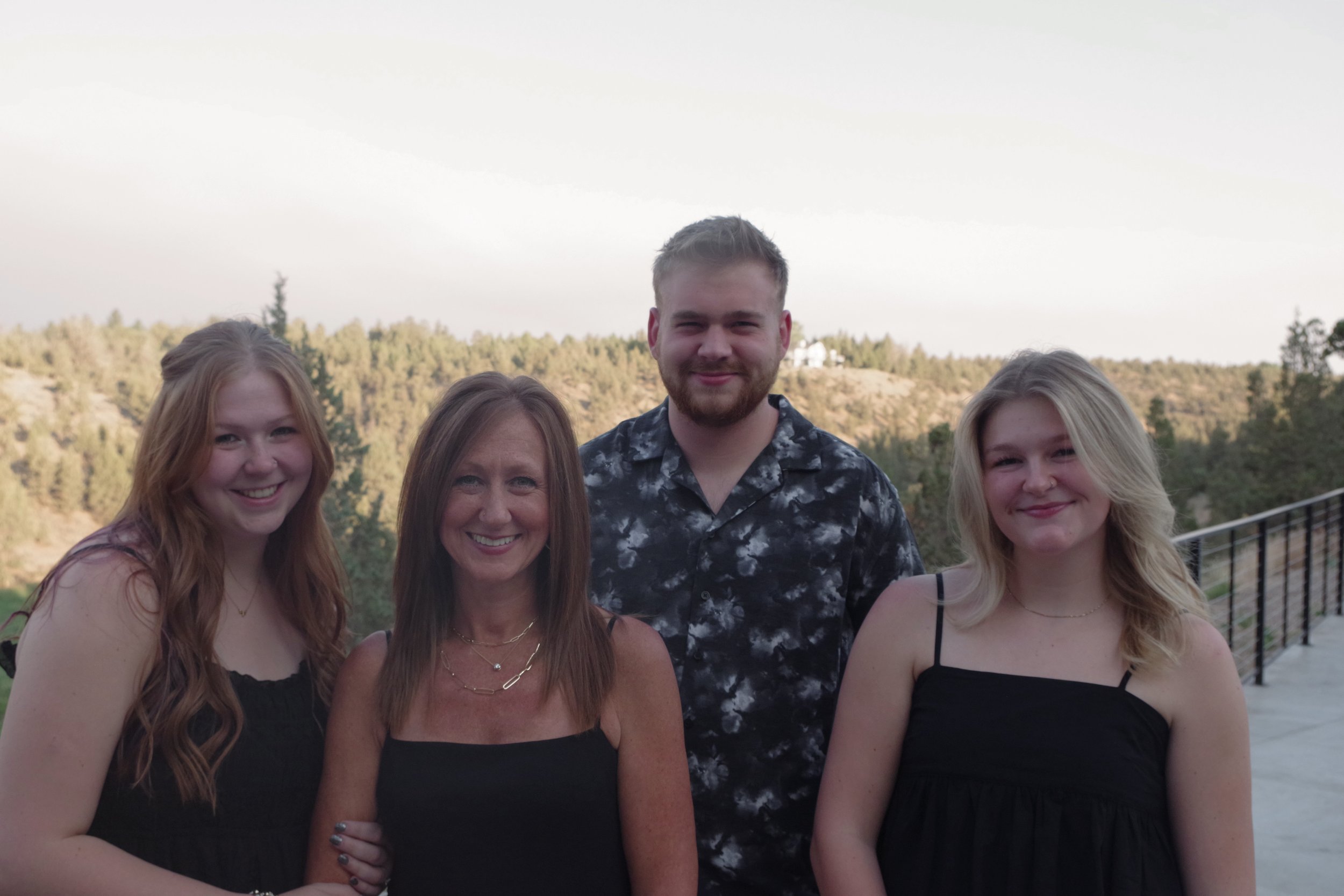 Group photo of five smiling adults standing outdoors with hills and trees in the background during daytime.