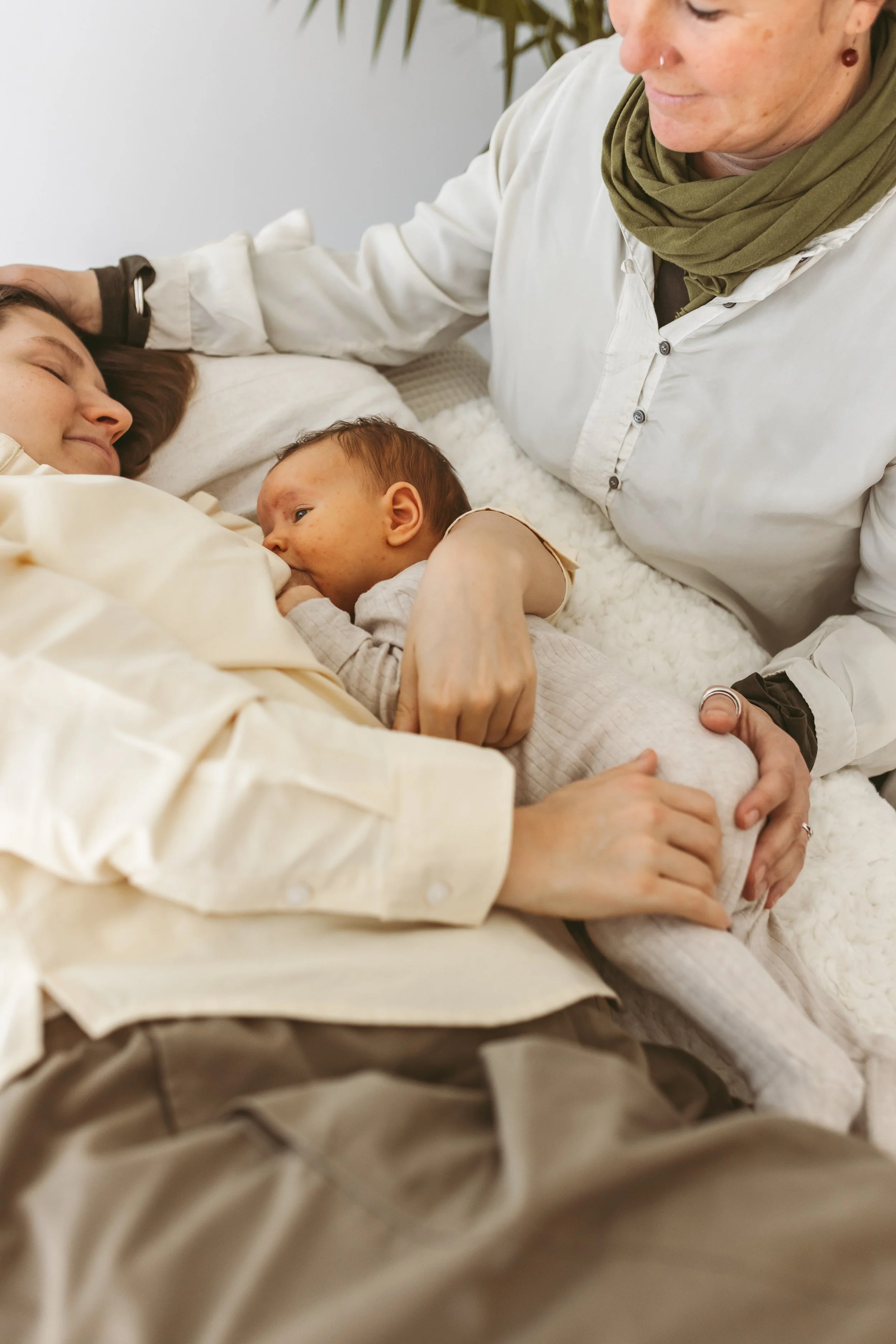 A woman is lying down, gently holding her newborn baby who is feeding at her breast. An older woman is sitting beside them, caressing the mother's head and holding the baby securely.