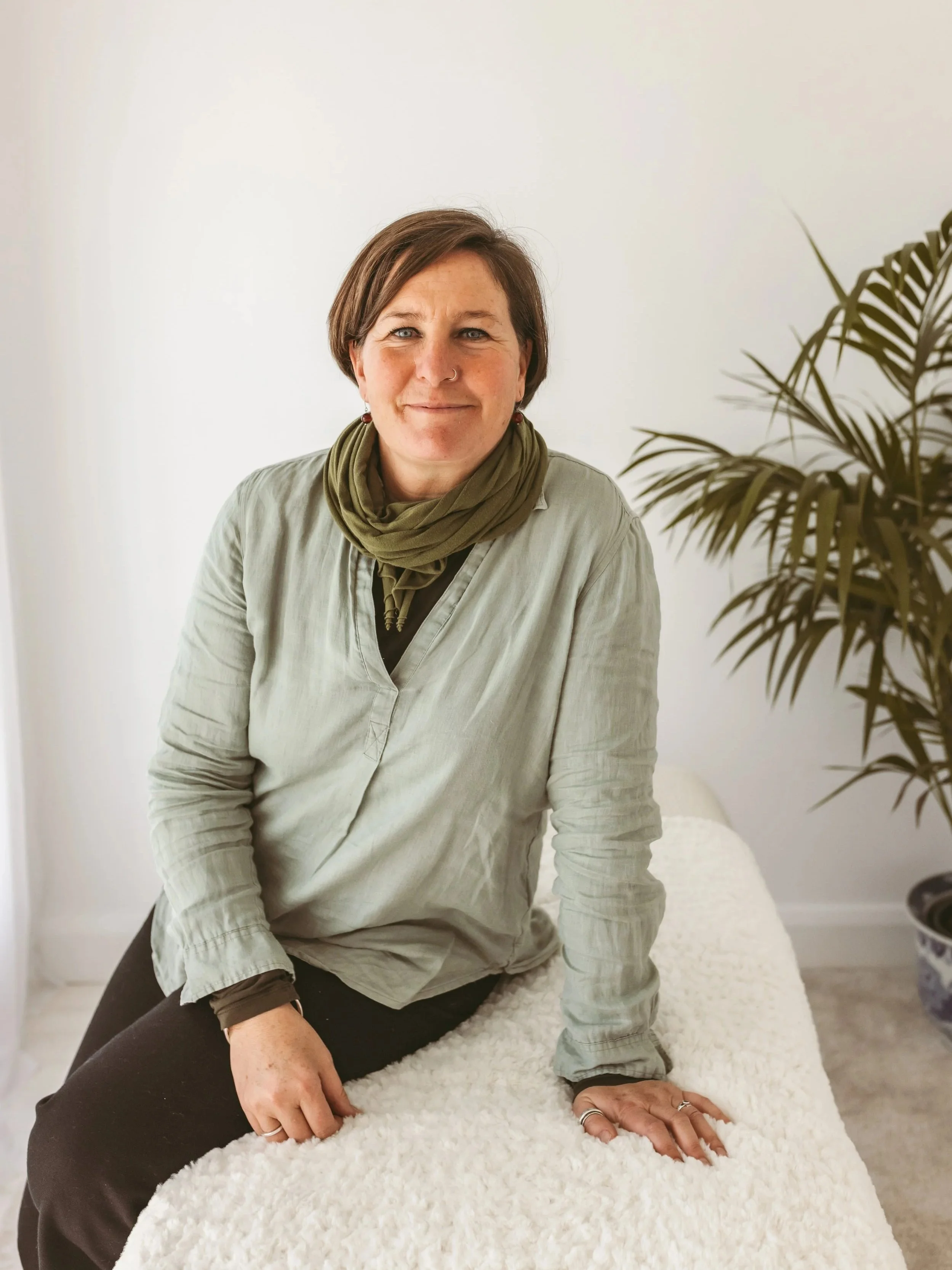 A woman with short brown hair, wearing a light-colored shirt and green scarf, sitting on a fluffy white surface indoors, with a plant in the background.