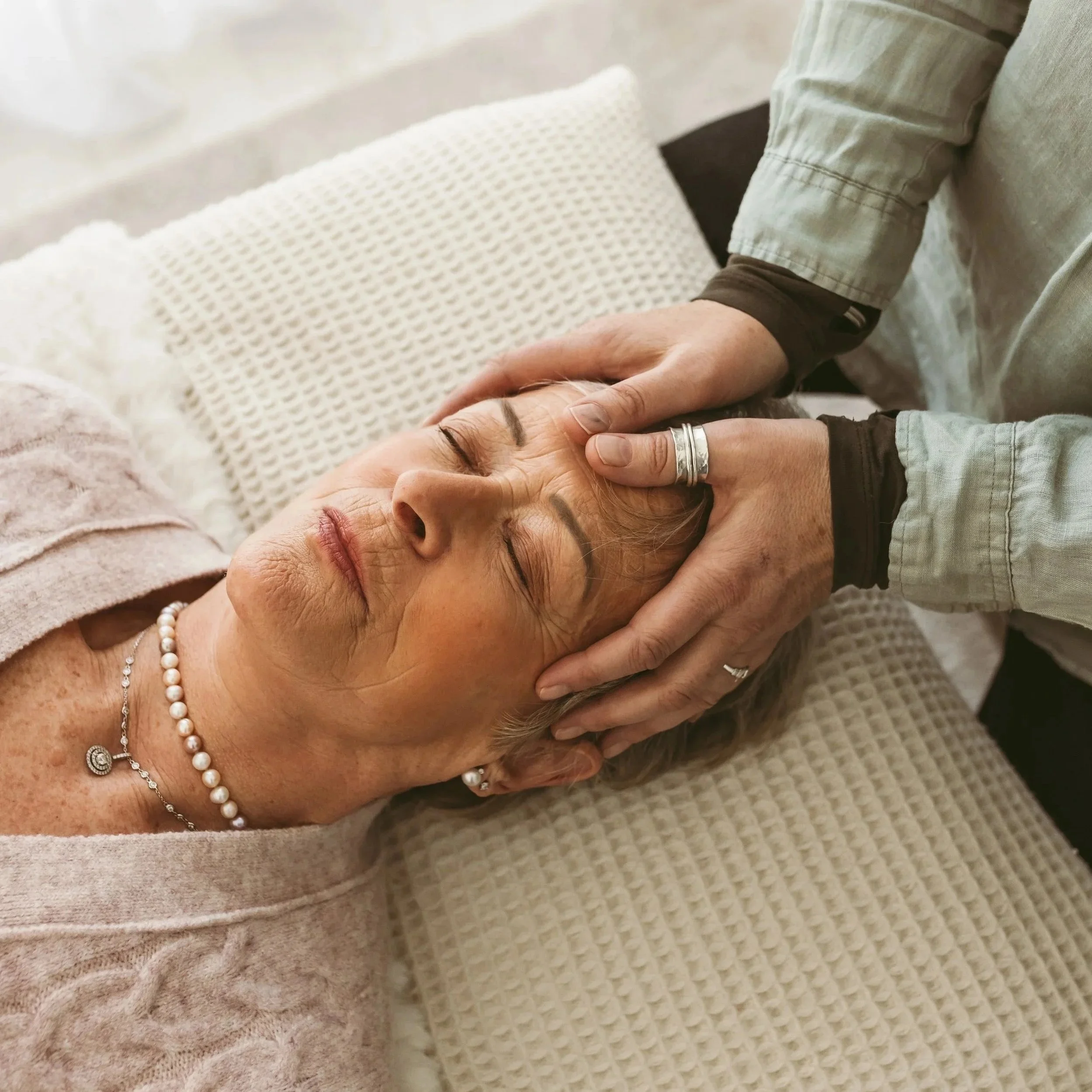 A person with a bracelet and rings gently holding the face of an elderly woman, who is lying down on a textured white blanket.