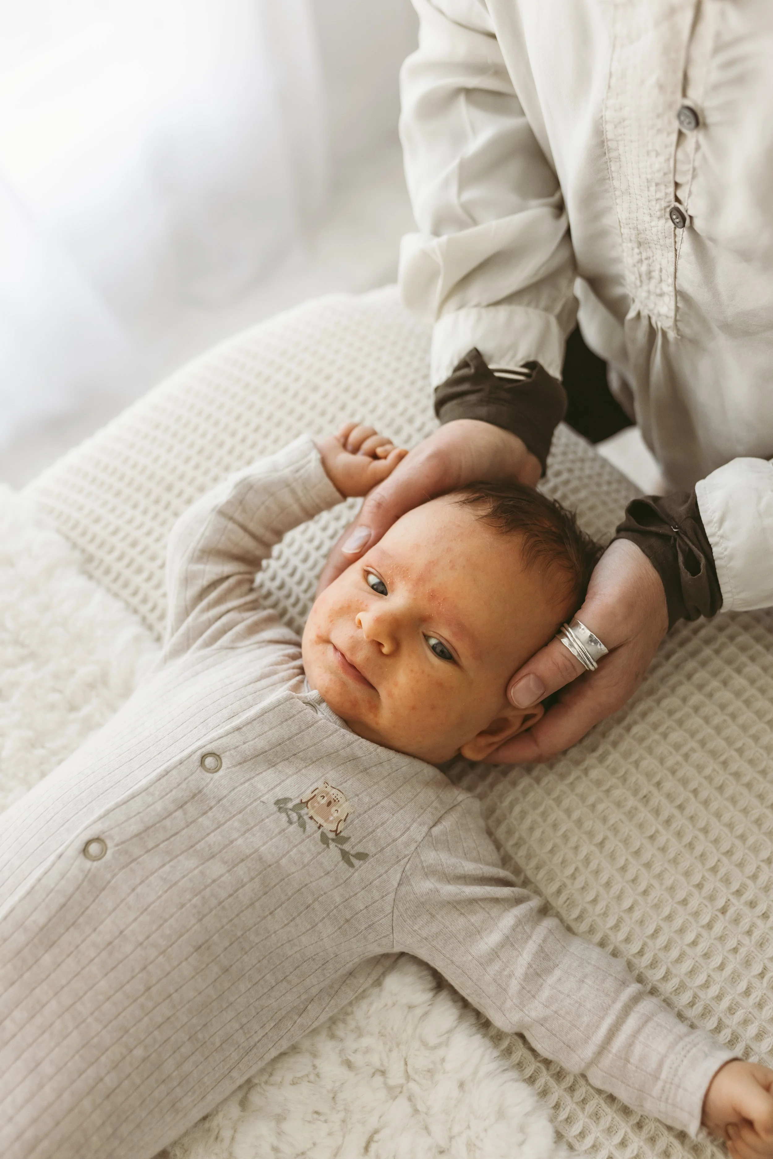 A young boy lying on a plush cream-colored rug, with a textured blanket and a person hand supporting his head, inside a cozy indoor setting.