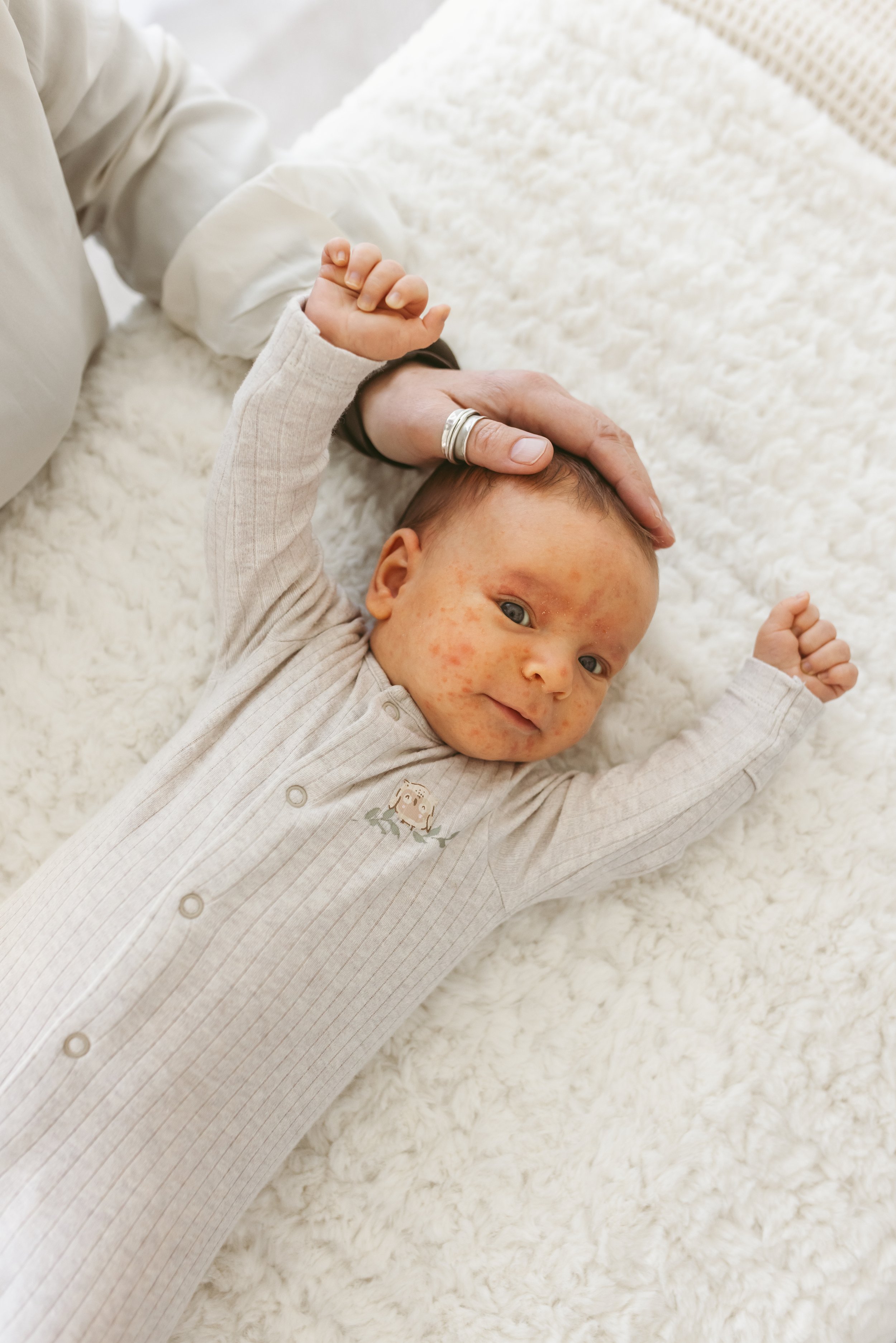 A baby lying on a white fluffy blanket, holding an adult's hand and looking at the camera.