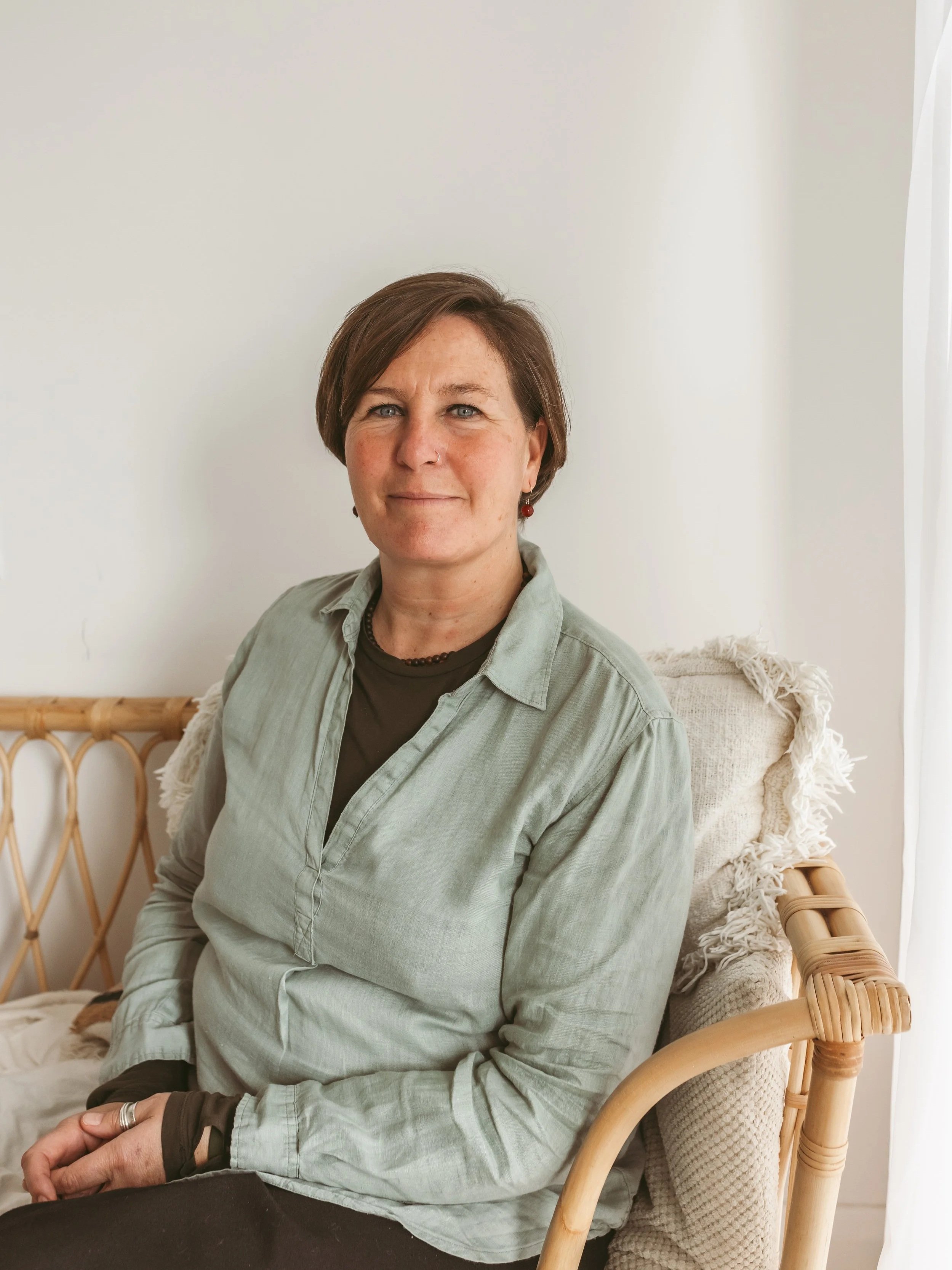 A woman with short brown hair, wearing a light gray button-up shirt and black pants, sitting on a wicker chair with a beige cushion. She has a small nose ring and is wearing earrings and a bracelet, with her hands clasped on her lap. She is in a room with white walls and a window with sheer curtains.