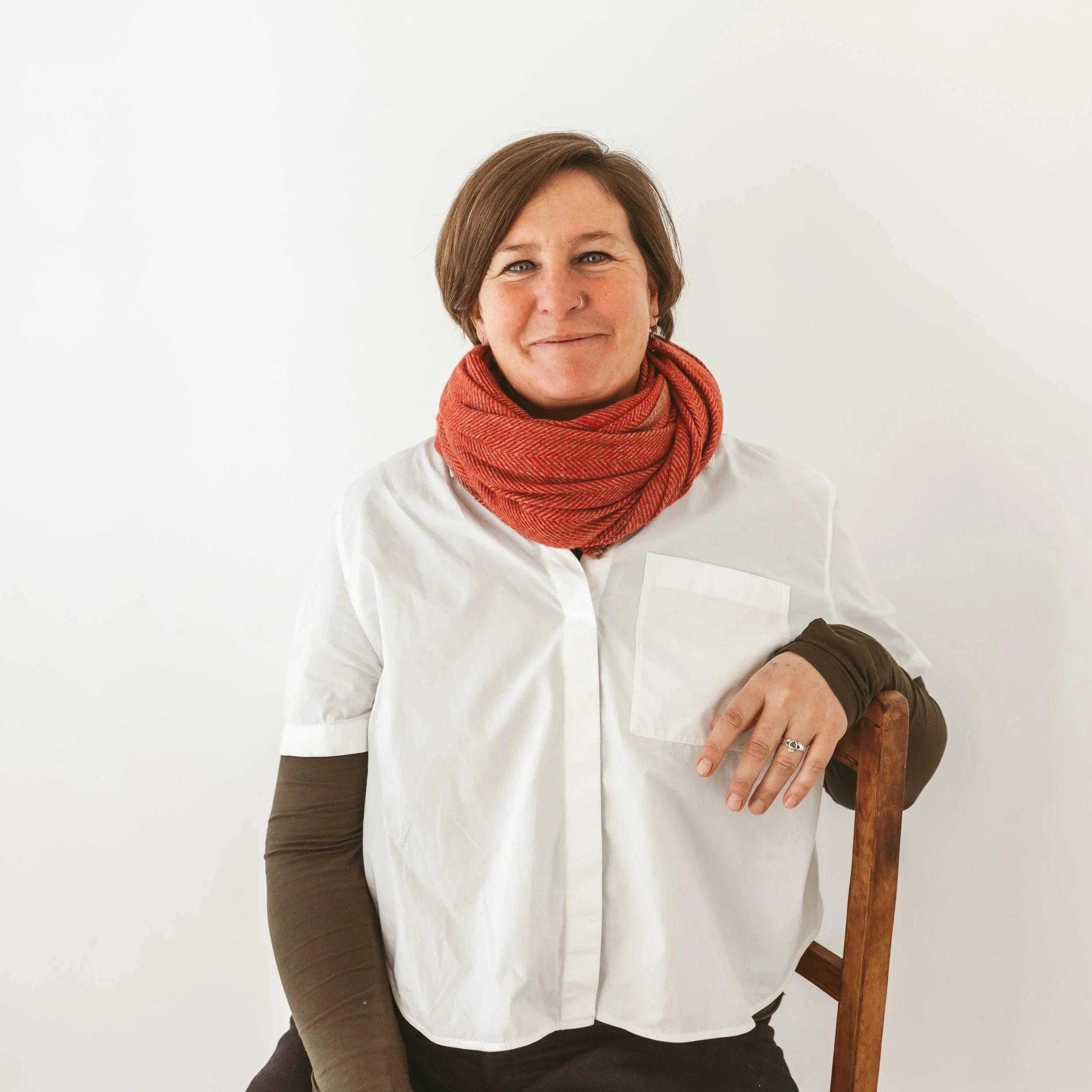 A woman with short brown hair and a nose ring, wearing a white shirt and a red scarf, sitting on a wooden chair, smiling, against a plain white wall.