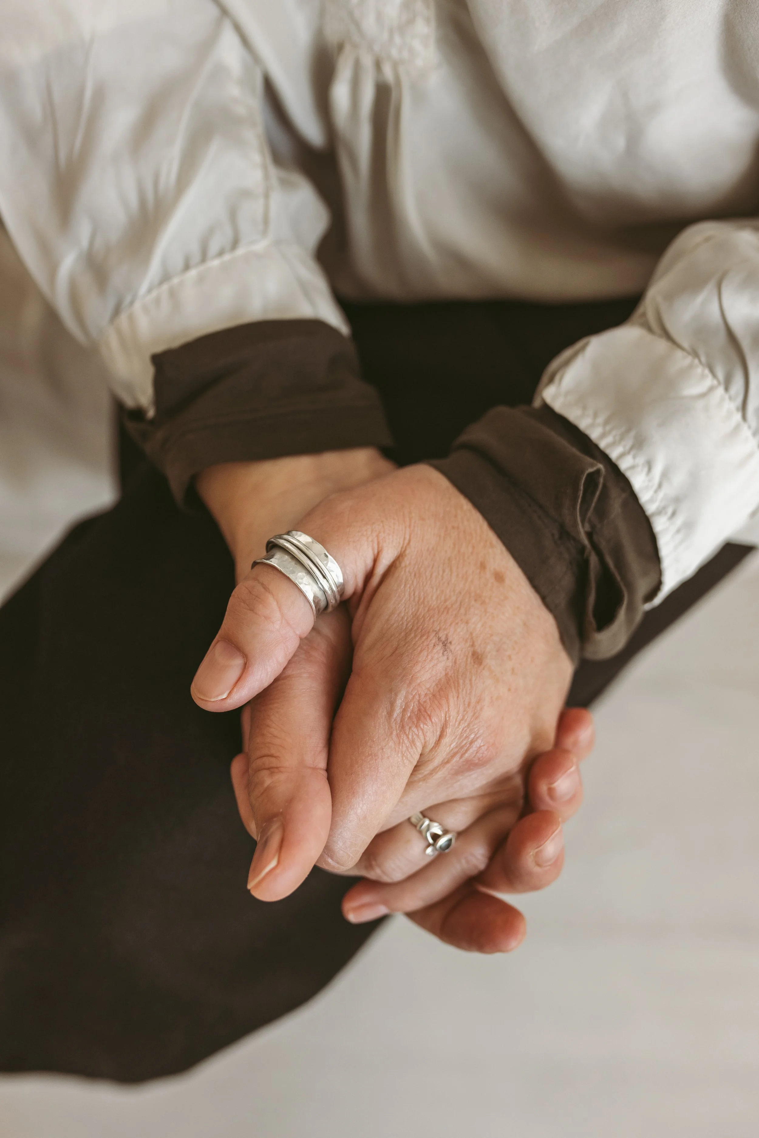 Close-up of two people holding hands with wedding bands, one wearing a white puffy sleeve jacket and the other a dark sleeve shirt.