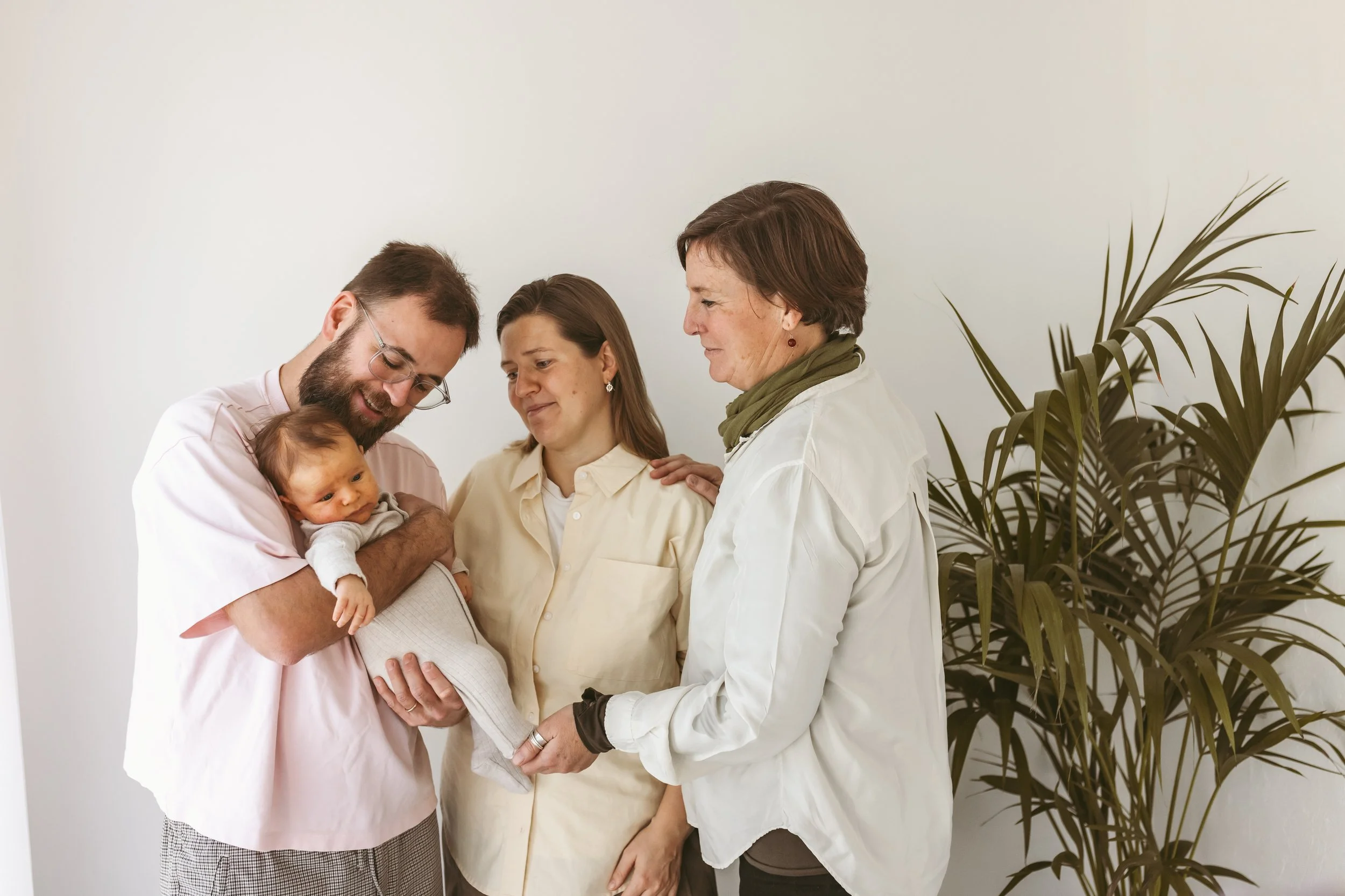 Family gathering with three women and a man holding a baby, standing near a large plant against a plain white wall.