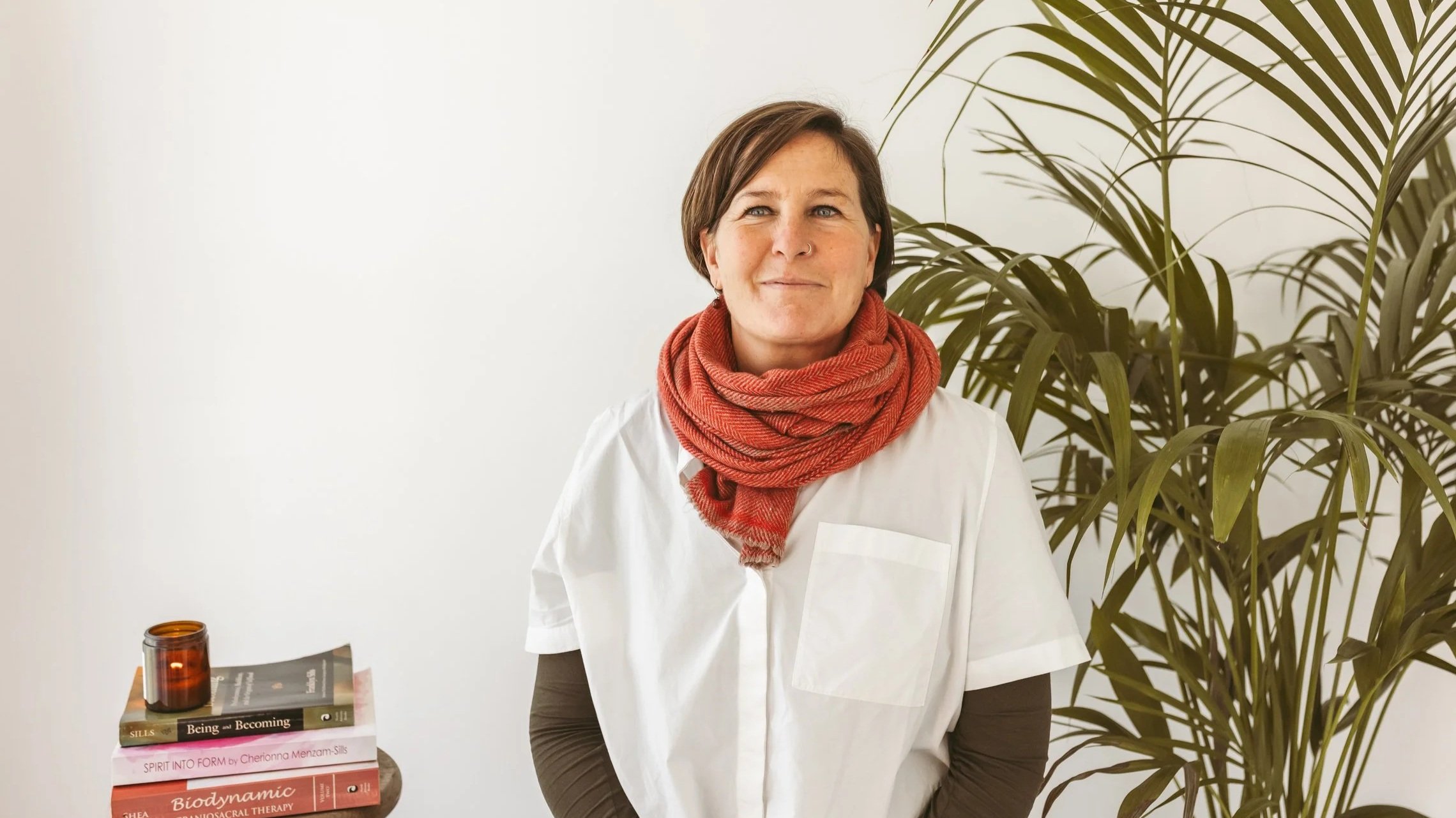A woman with short brown hair and blue eyes, wearing a red scarf, white shirt, and gray sleeves, standing near a large green indoor plant, with a stack of books and a candle on a table to her left.