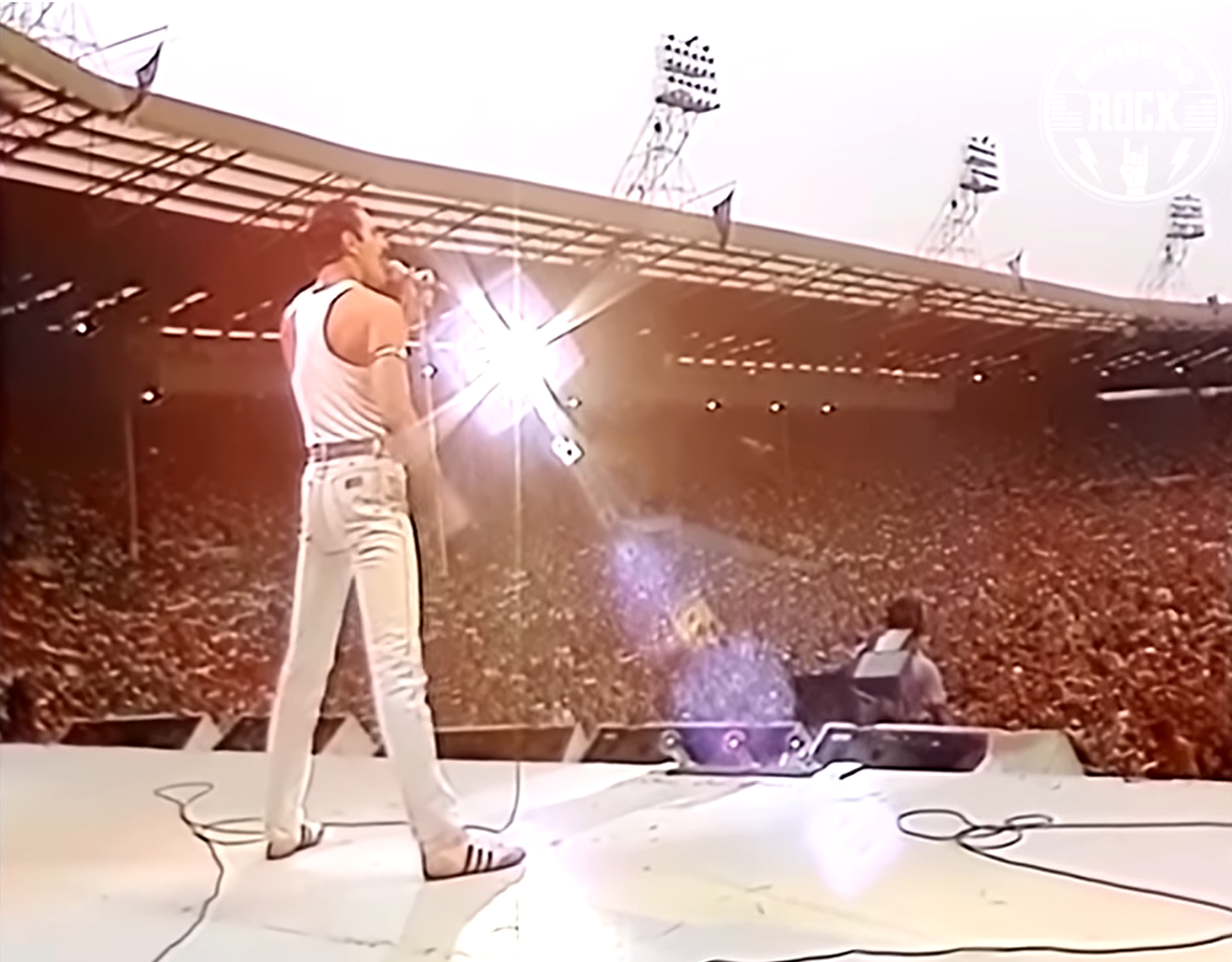 A singer performer on stage with a microphone in hand, facing a large crowd at an outdoor concert stadium during daytime.